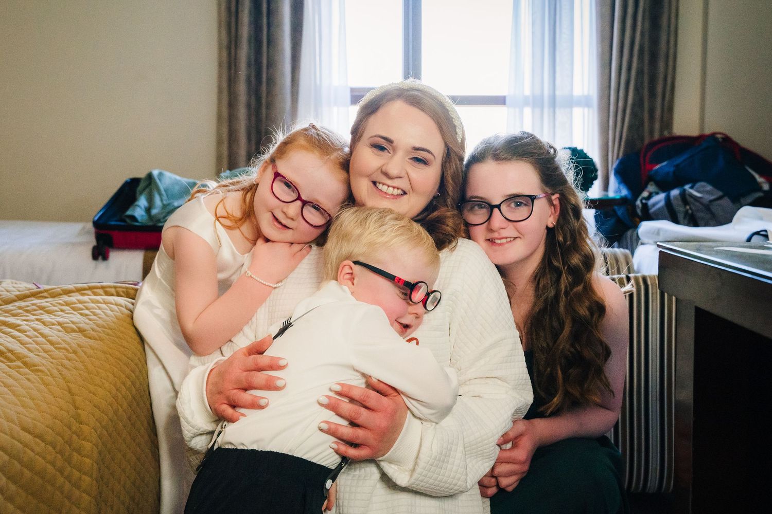 Four people wearing white sweaters share a warm embrace while sitting on a gold colored couch in a bright room.  Wedding PHOTOGRAPHER Trim C