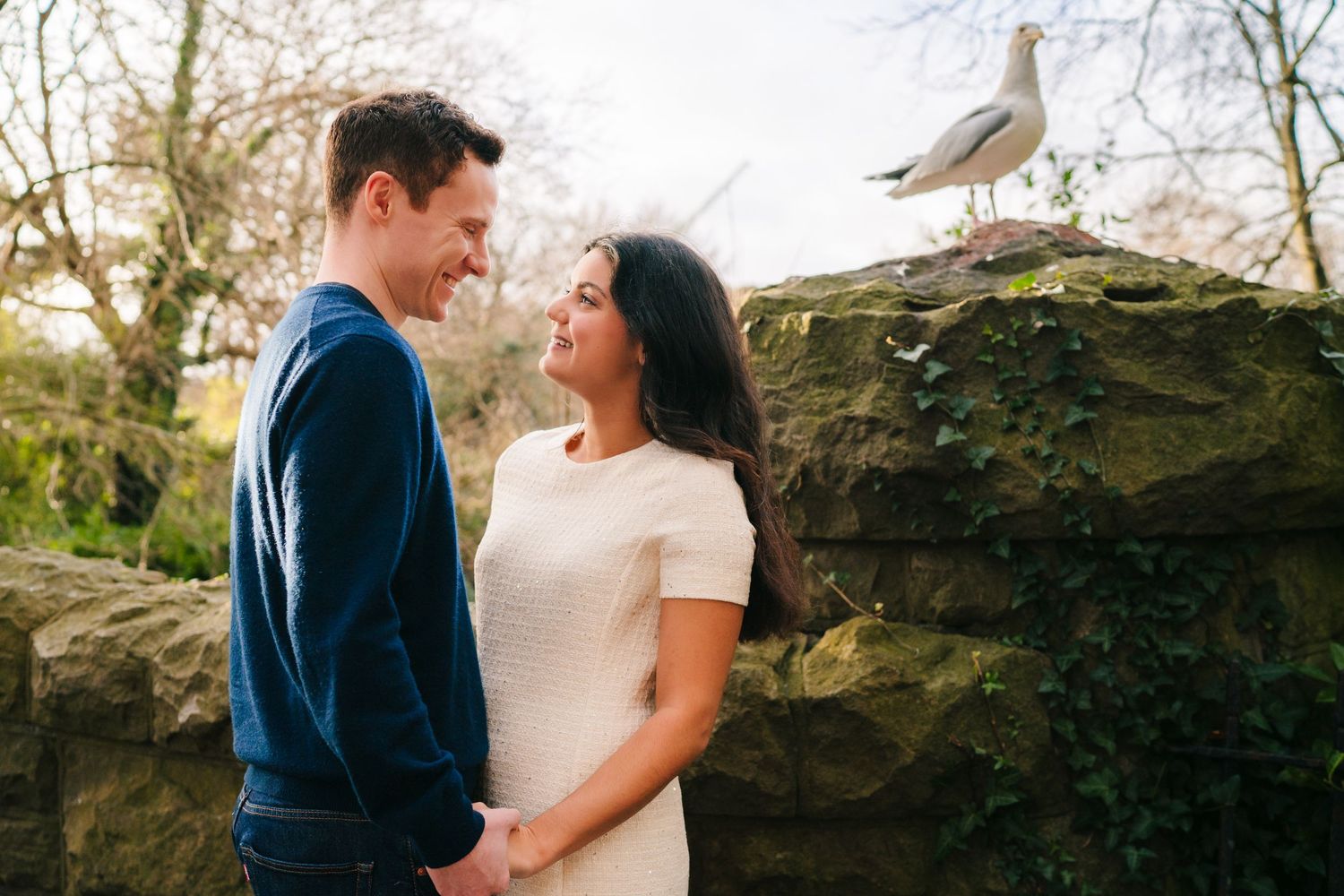 A couple in wedding attire stands among large moss-covered rocks in an outdoor garden setting.Surprise proposal dublin photography