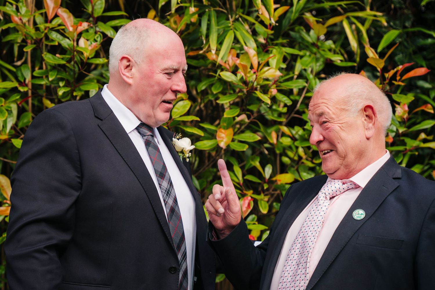 Two men in dark suits engaged in animated conversation against a garden background. Portmarnock Hotel Wedding PHOTOGRAPHER Dublin
