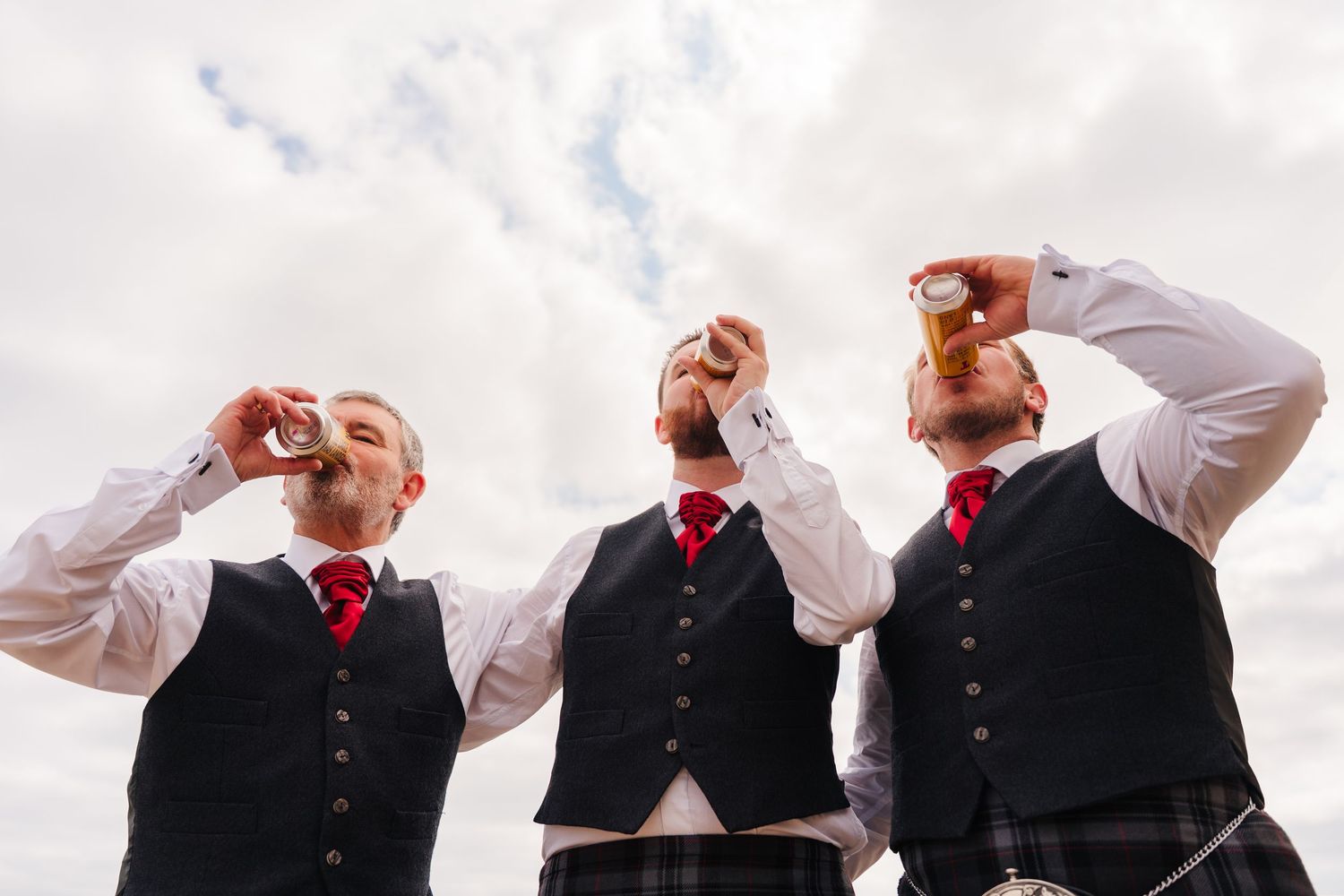 Three groomsmen in matching black vests and red ties drink from beer bottles against a cloudy sky. Portmarnock Hotel Wedding PHOTOGRAPHER Du