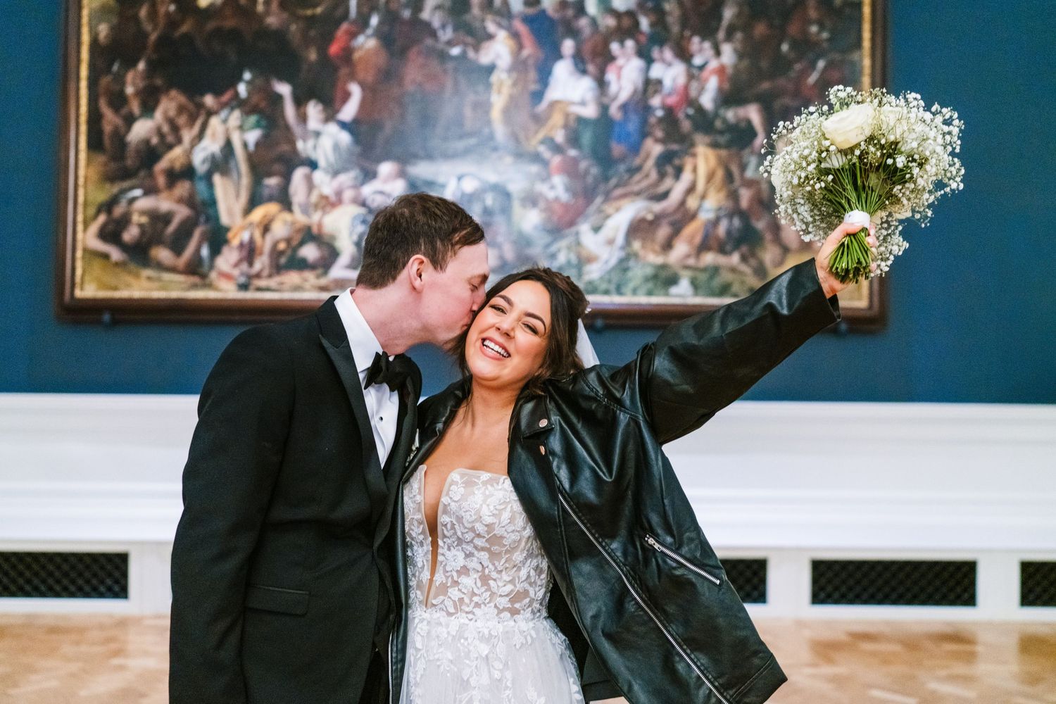 A bride wearing a leather jacket holds up her bouquet while celebrating in an elegant room. Wedding PHOTOGRAPHER Dublin