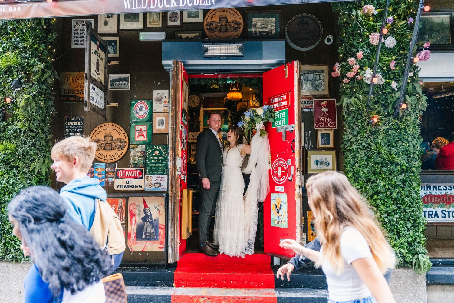 Wedding couple stands in a red-carpeted doorway of a charming storefront decorated with posters and framed art. PHOTOGRAPHER Dublin Ireland
