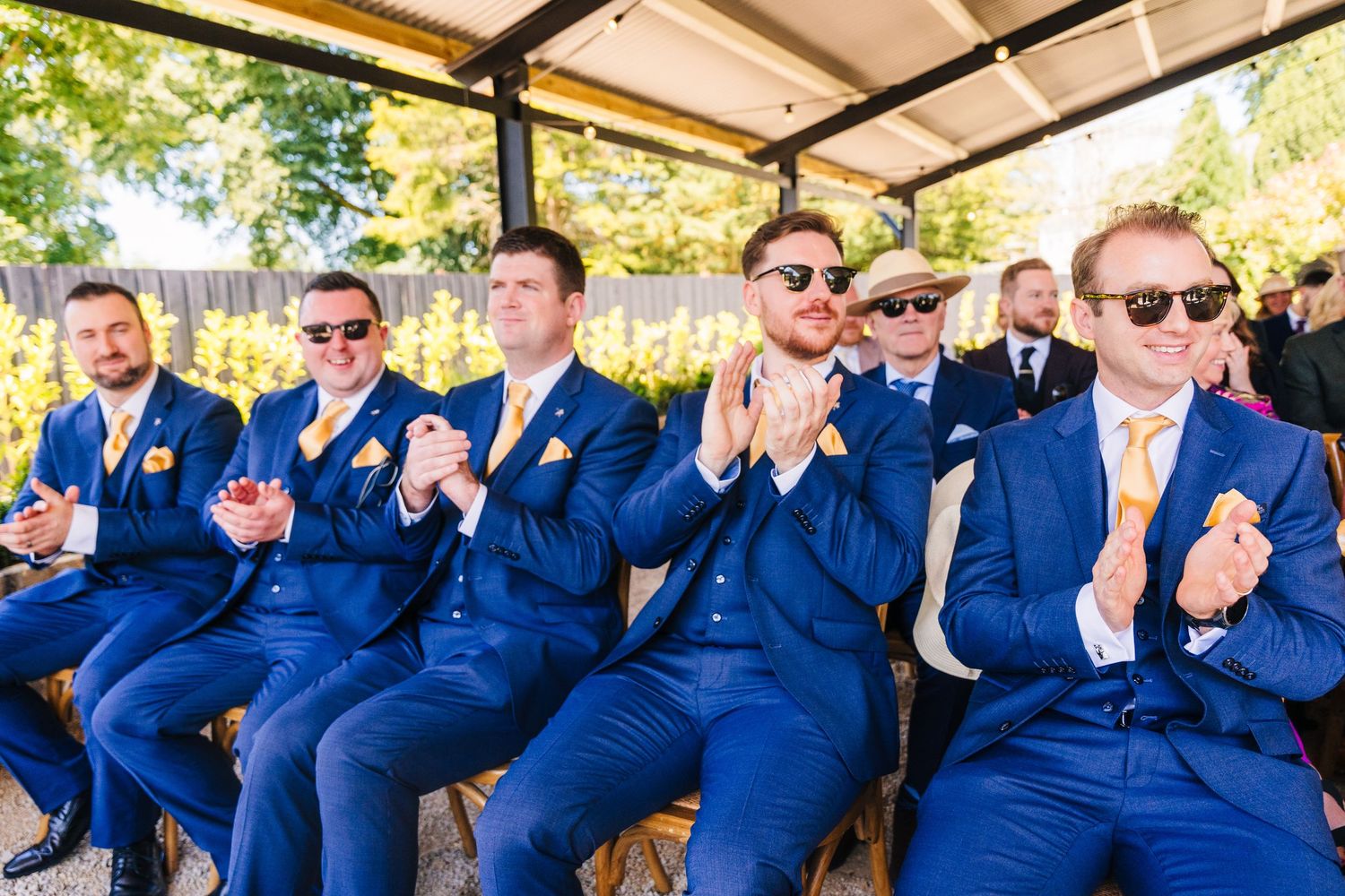 Poulaphouca Wedding photographer wicklow  Groomsmen in matching navy blue suits wearing sunglasses clap while seated outdoors.
