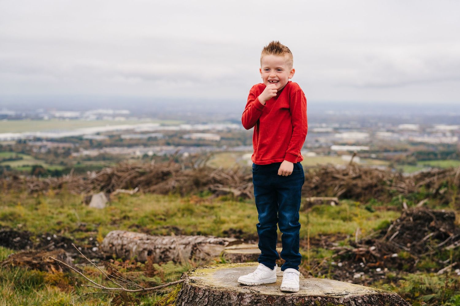 Family Photographer Dublin Ireland A small child in a red sweater stands on rocks overlooking a vast cityscape on a cloudy day.