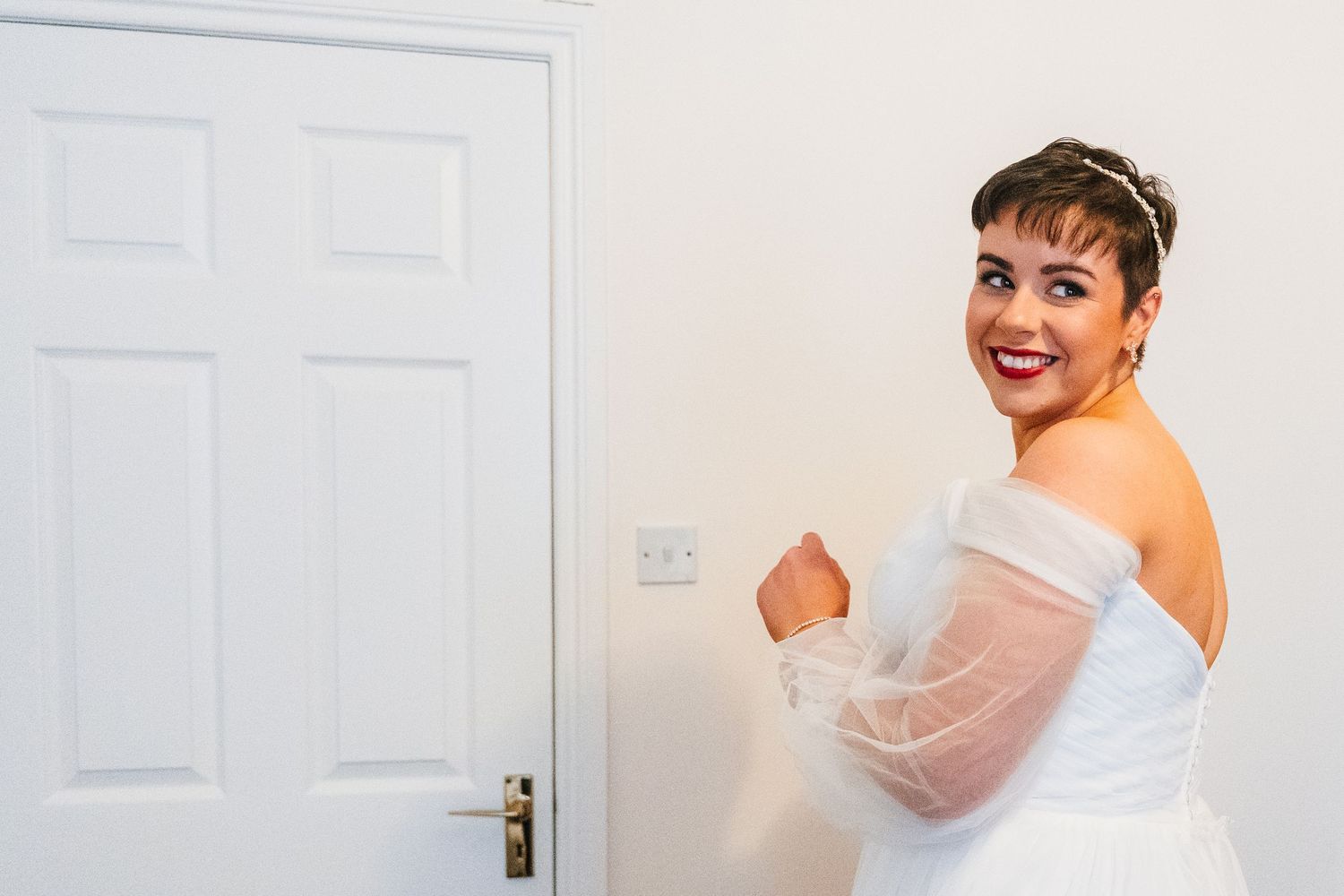 A joyful bride stands near a white door wearing an off-shoulder wedding dress with sheer sleeves. Millhouse slane wedding photographer dubli
