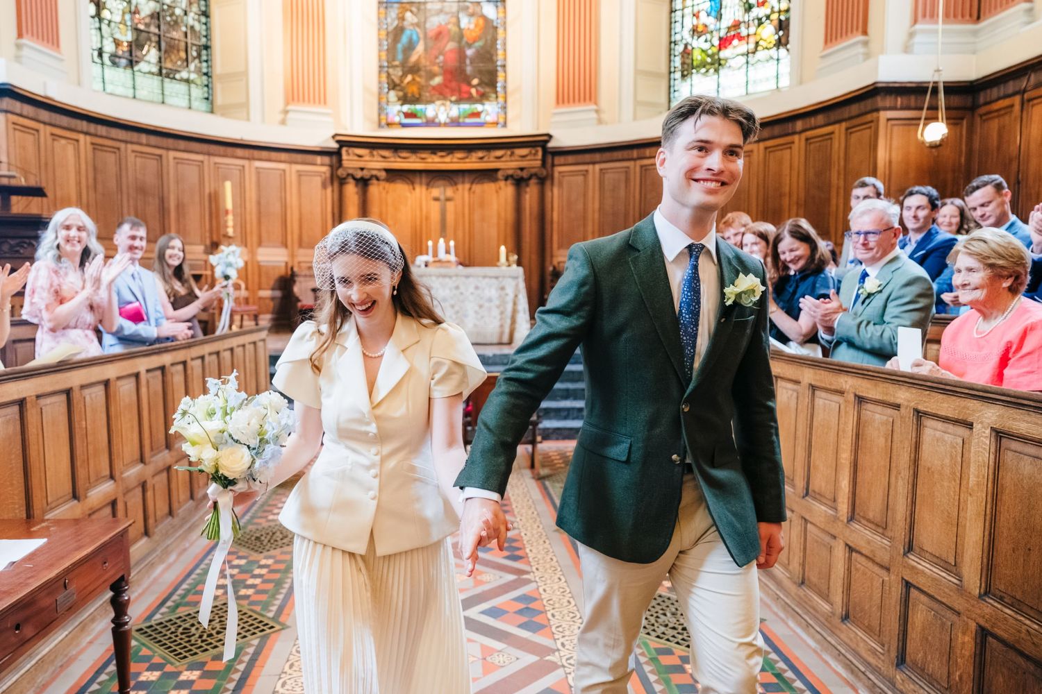Joyful newlyweds walk down church aisle, bride in cream suit holding bouquet, groom in green blazer, guests applauding.