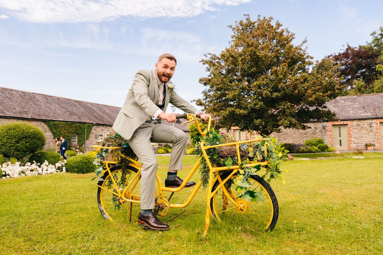 Wedding photographer Dublin Man in gray suit posing on a yellow tandem bicycle decorated with flowers and greenery in a garden setting.