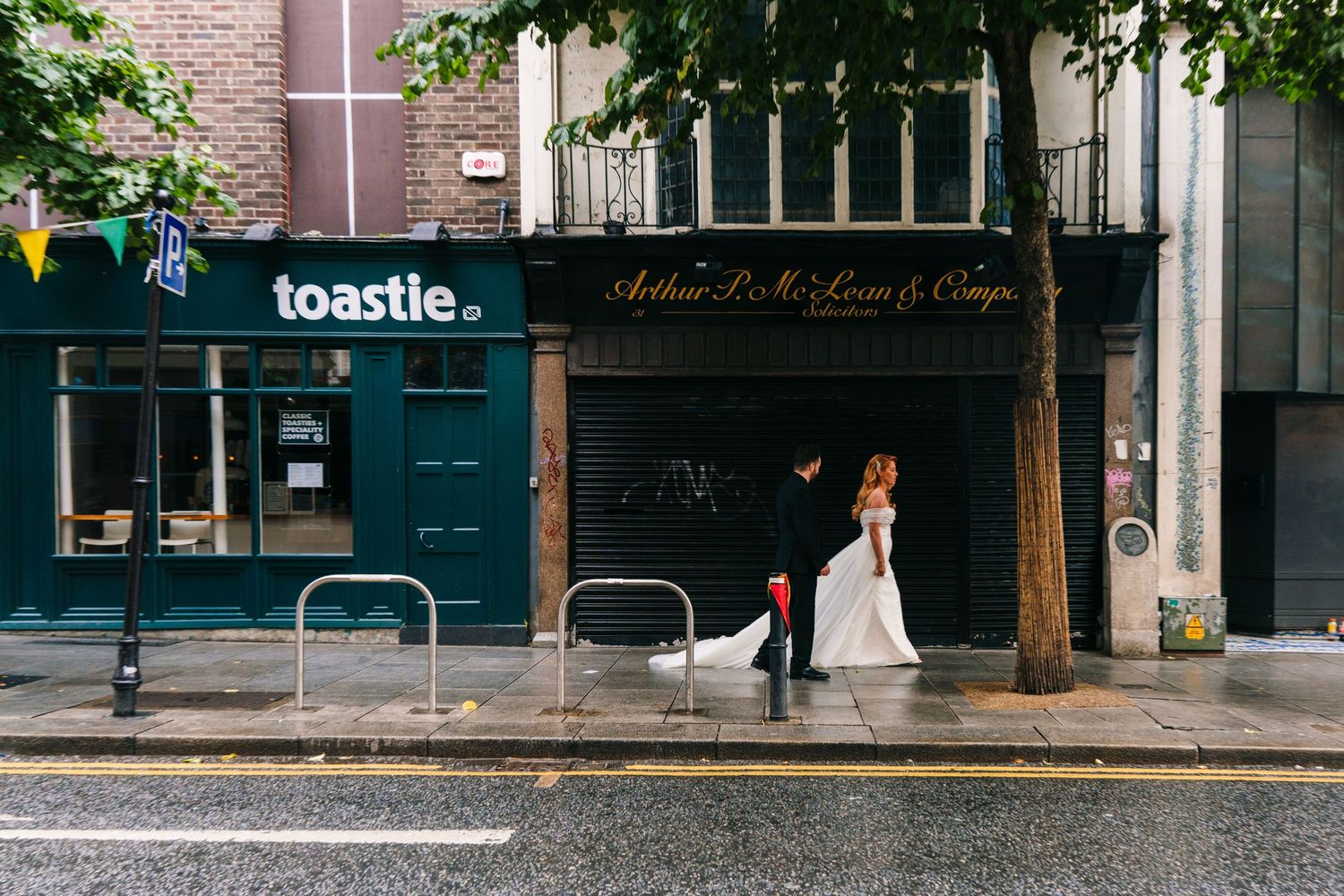 A wedding couple poses on a city street with storefronts and trees in the background.  city hall dublin wedding photos