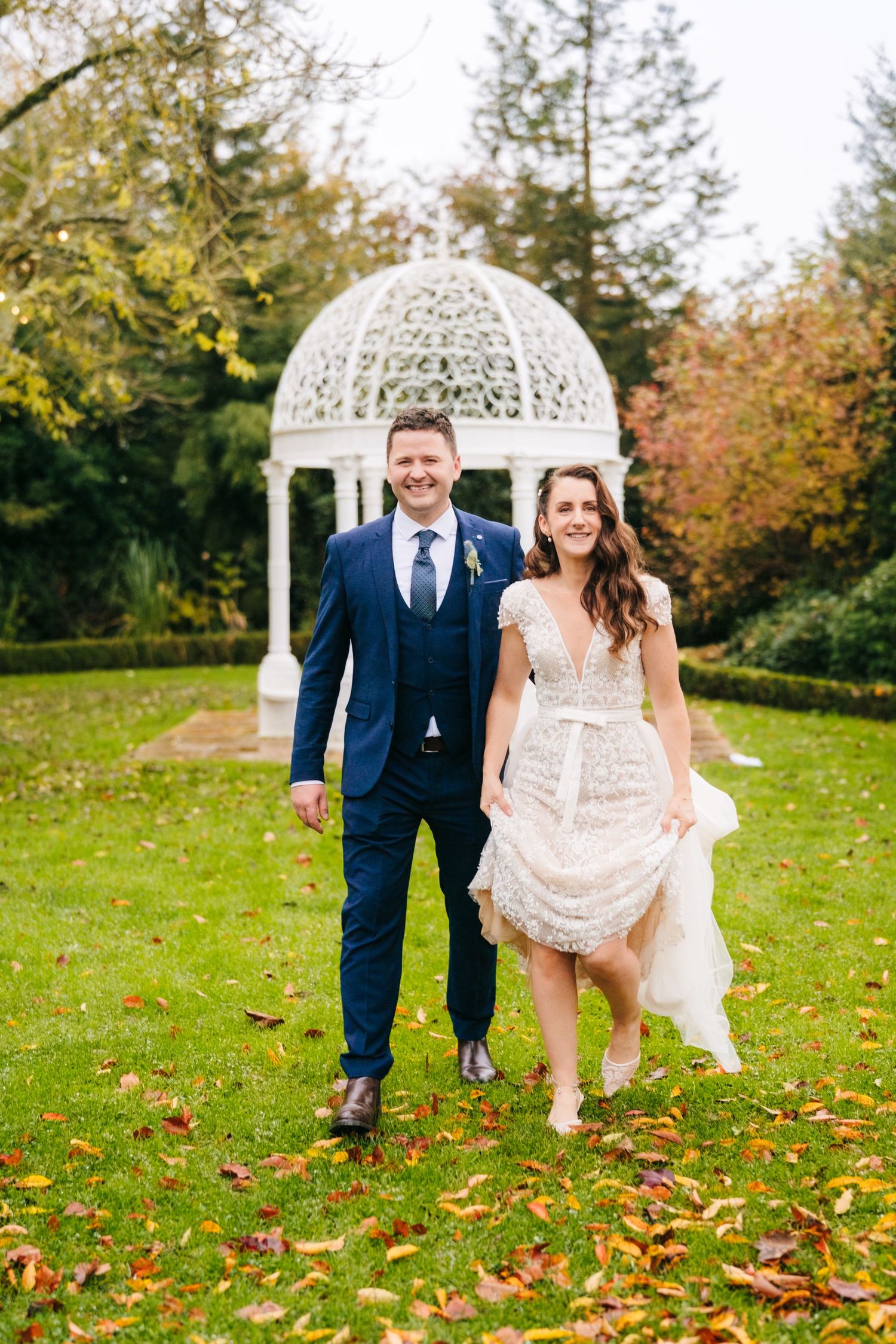 Couple walks through fallen leaves near a white garden gazebo on their wedding day.