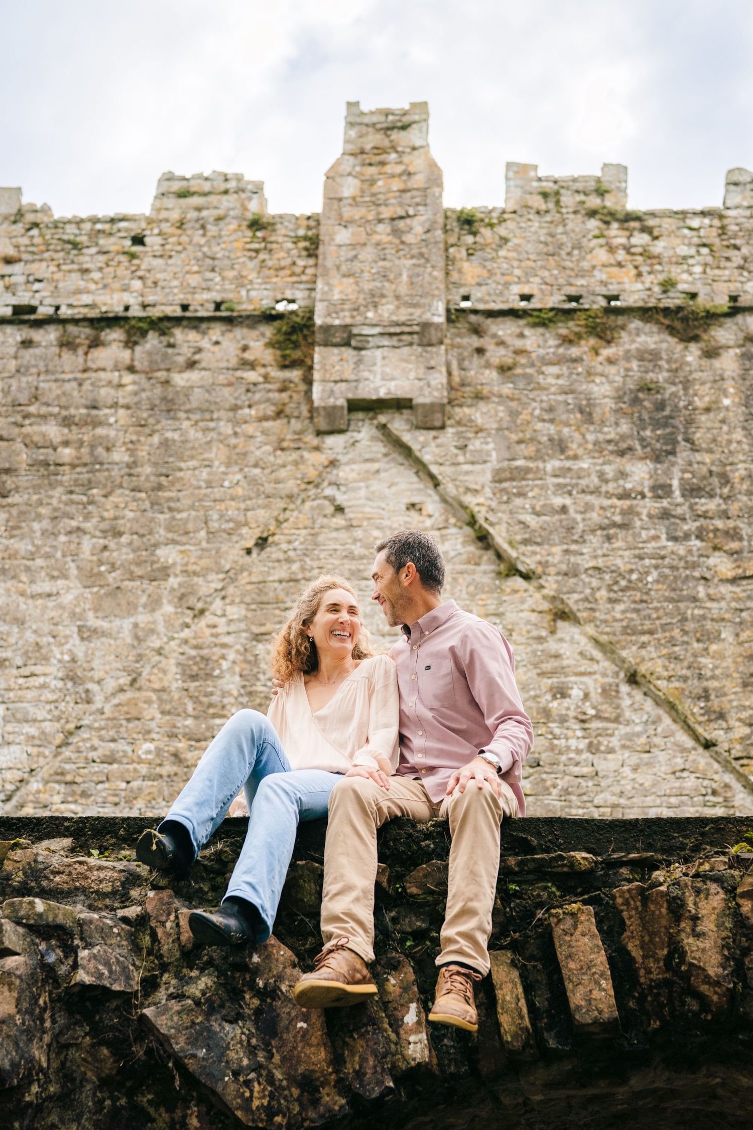 A couple sits together on an ancient stone wall at a historic castle during an engagement photo session. BEctive abbey family photographer d