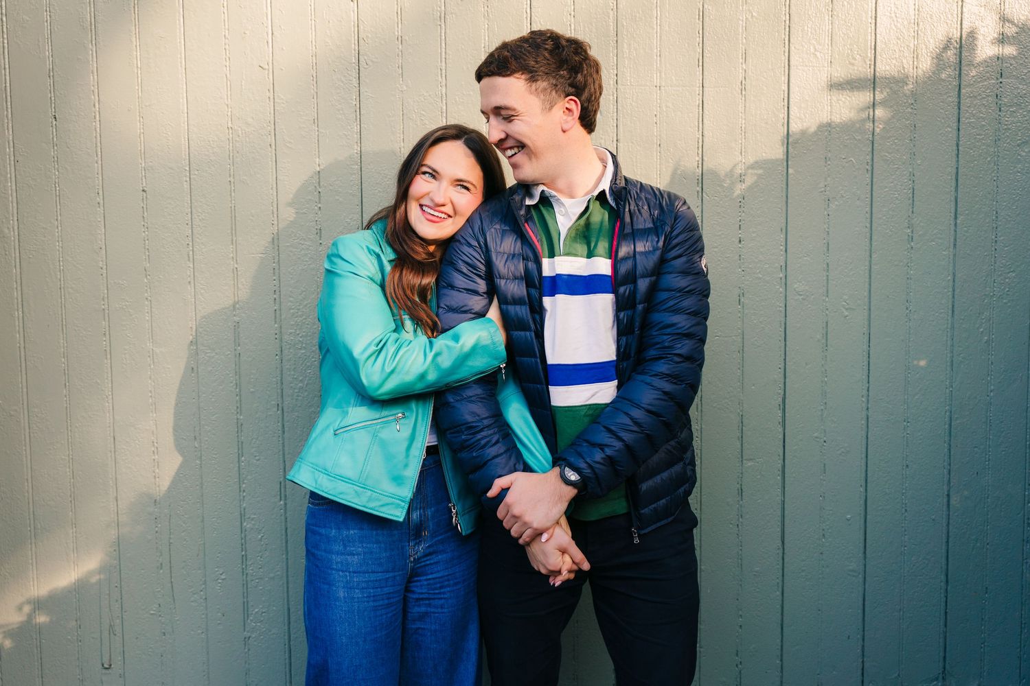 engagement photographer dublin A couple in blue winter jackets laughing together while leaning against a gray wall.
