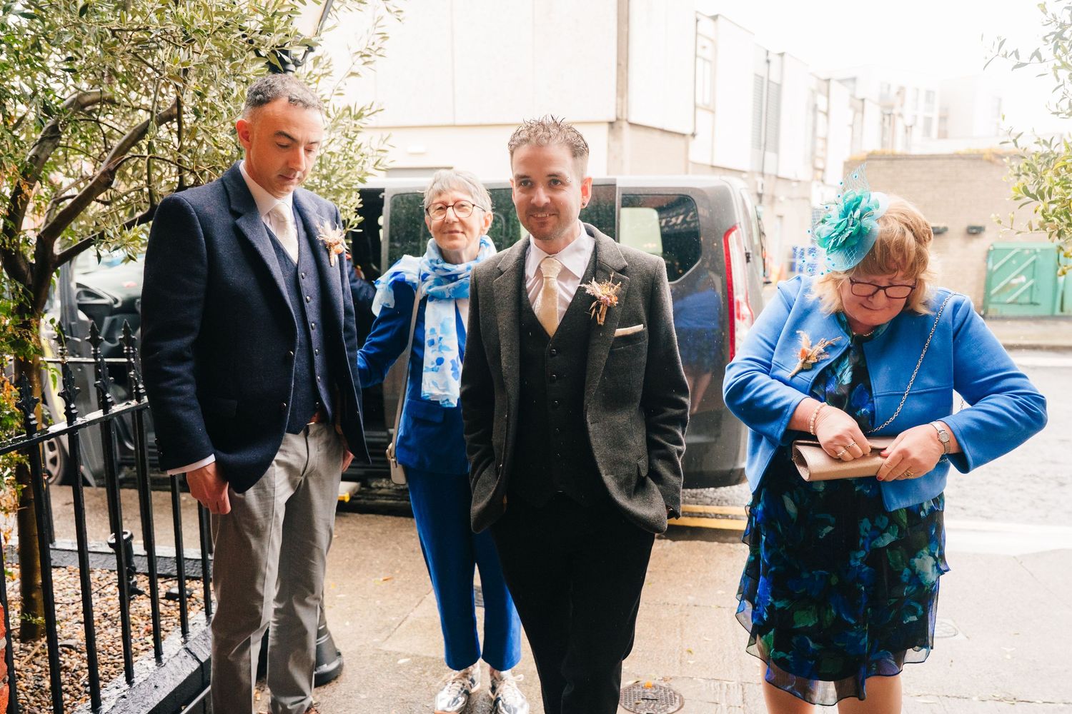 Wedding party members gather outside near a vehicle on a sunny day.