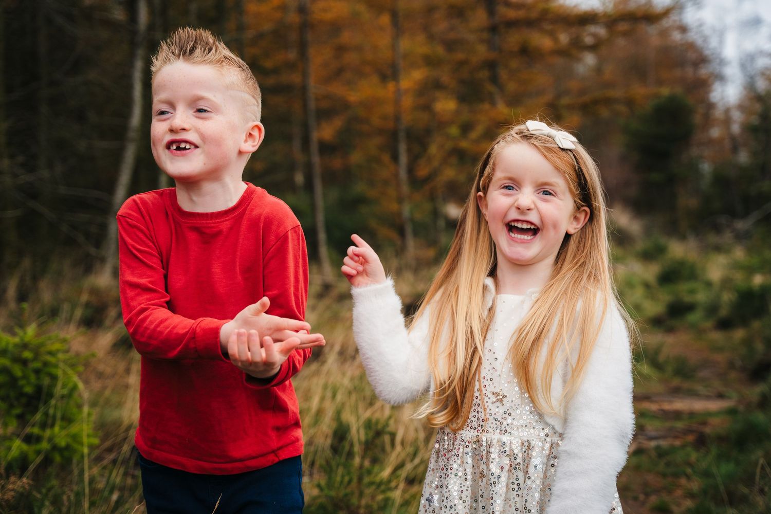 Two children laugh together outdoors during autumn wearing red and white sweaters in front of fall foliage. family PHOTOGRAPHER Dublin