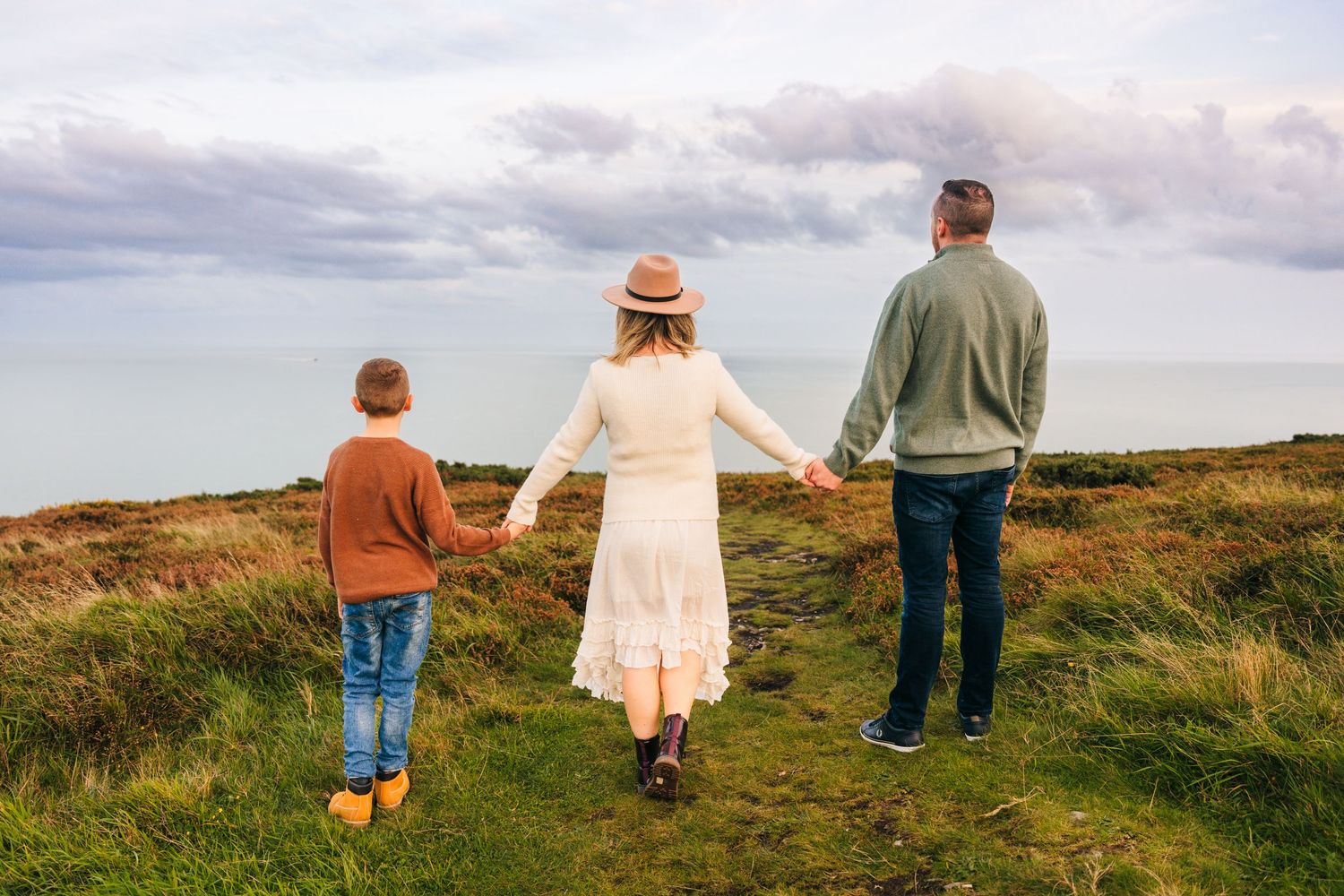 Family walking hand in hand along scenic coastal path on sunny day with cloudy sky and rolling hills. Family PHOTOGRAPHER Dublin