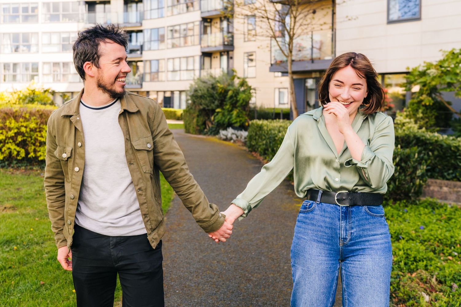 Happy couple holding hands while walking on a sidewalk path outside an apartment building on a sunny day.