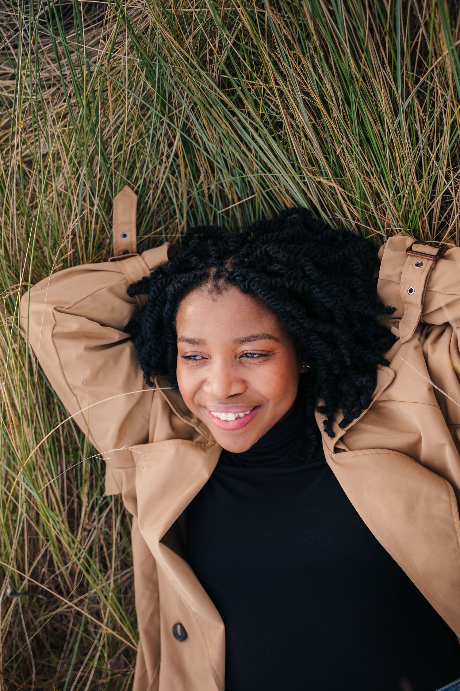 Person with natural curly hair lying in tall grass wearing a tan coat and black turtleneck while smiling. PHOTOGRAPHER Dublin ireland