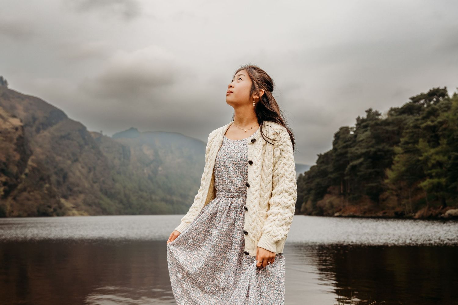 Person in a floral dress and cream cardigan stands peacefully by a mountain lake under moody skies. Family PHOTOGRAPHER Dublin