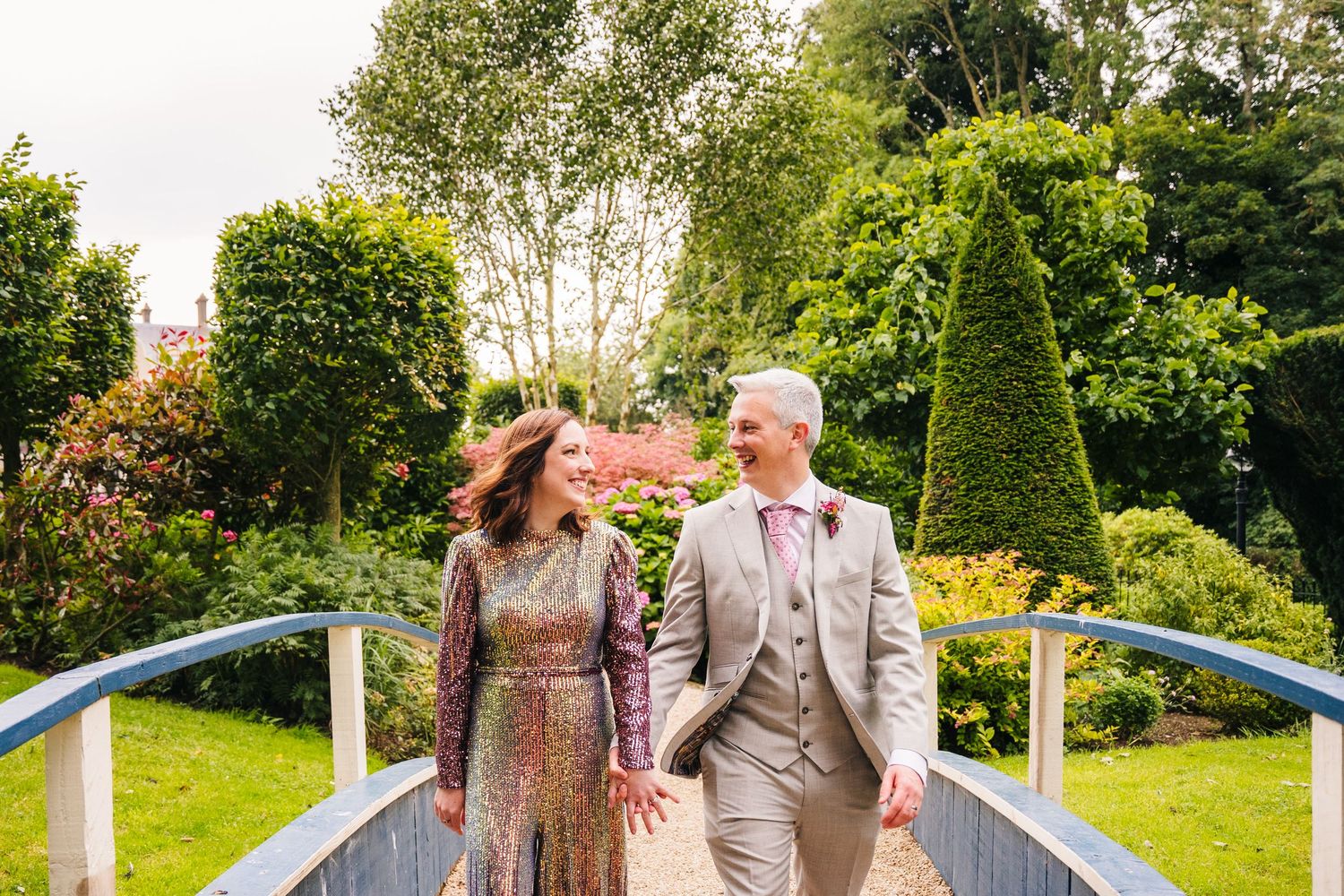 A couple walks together on a garden bridge surrounded by lush greenery and landscaped topiary bushes. Wedding PHOTOGRAPHER Dublin