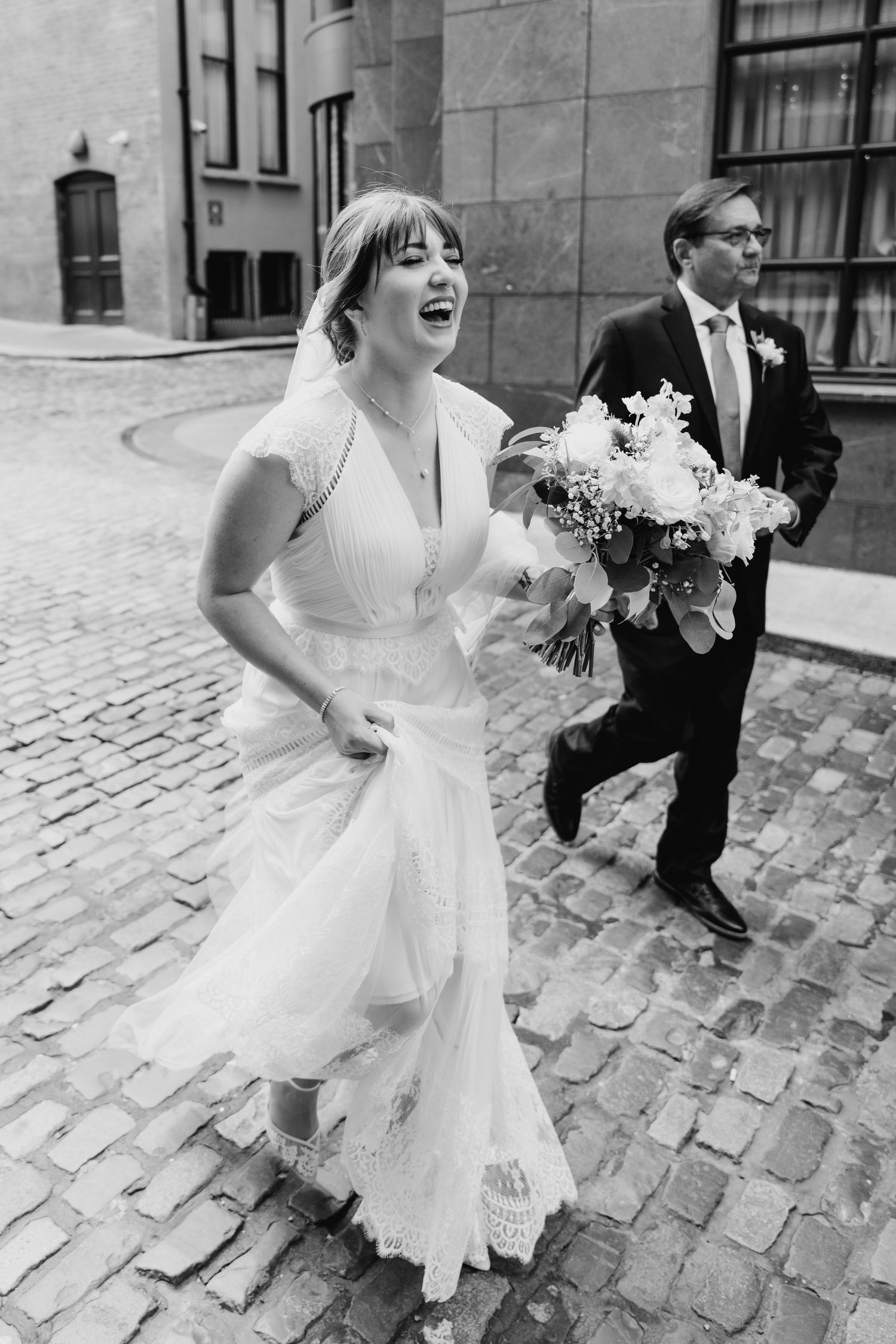 A candid black and white photo of a joyful wedding couple laughing while walking on cobblestone streets. Wedding PHOTOGRAPHER Dublin