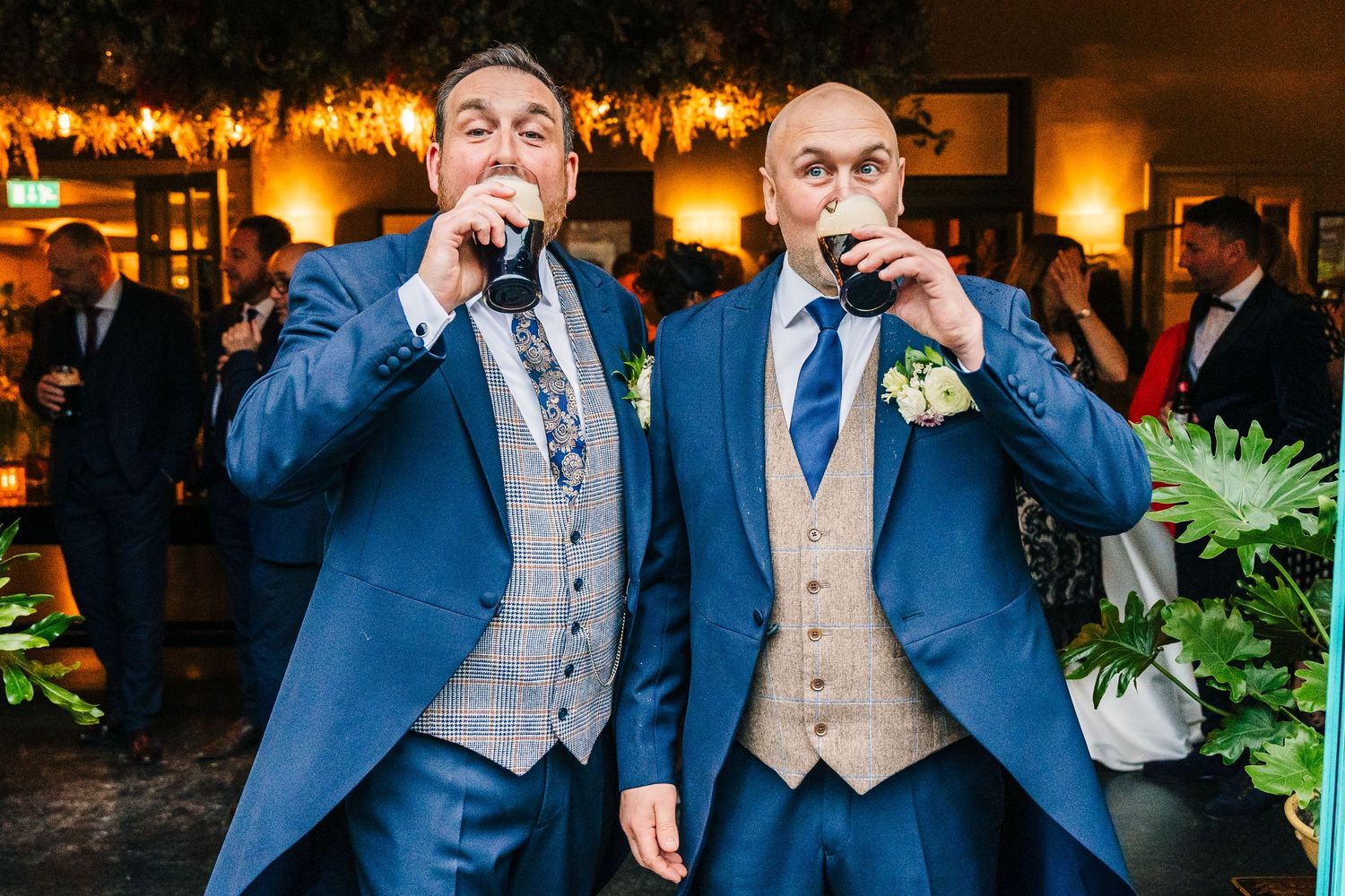 Two groomsmen in matching blue suits and gray vests share a celebratory drink at a wedding reception.