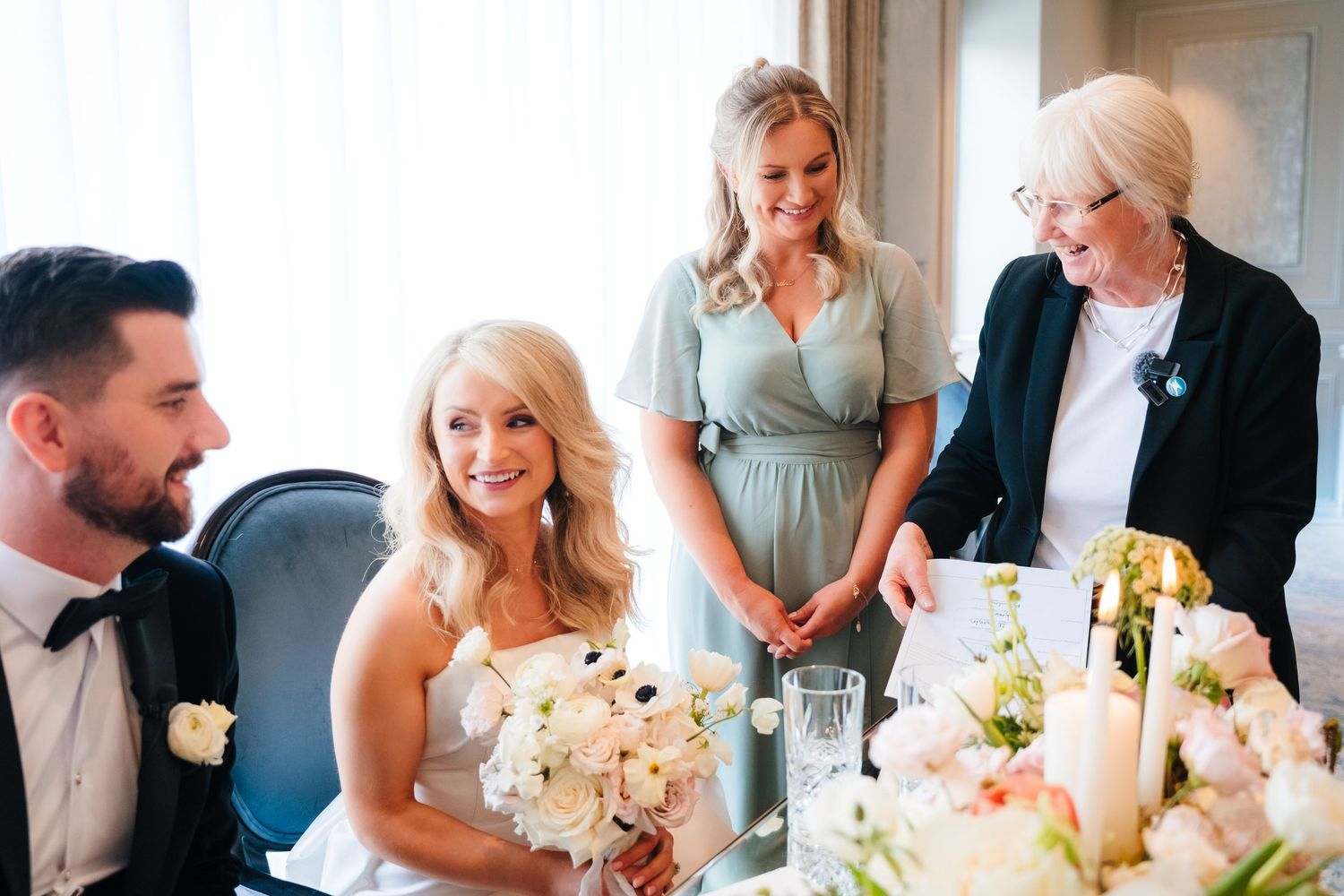Bride smiling at wedding reception table with bridesmaid and elderly guest, holding white floral bouquet.