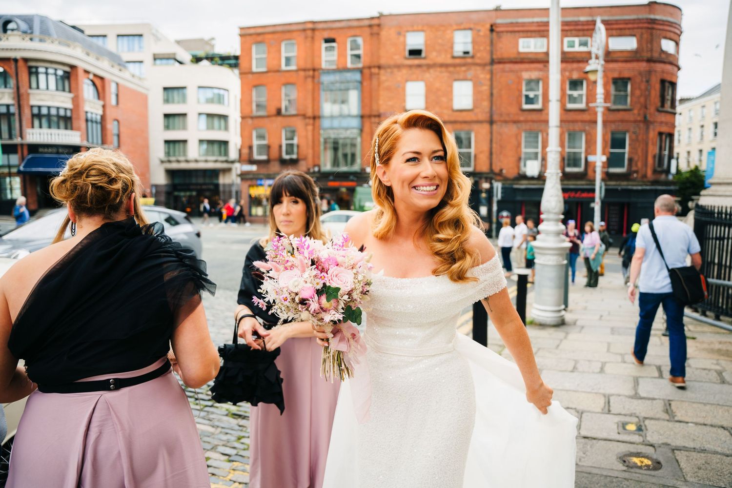 Wedding party wearing pink bridesmaid dresses and white gown pose together on cobblestone street near brick buildings.  city hall dublin wed