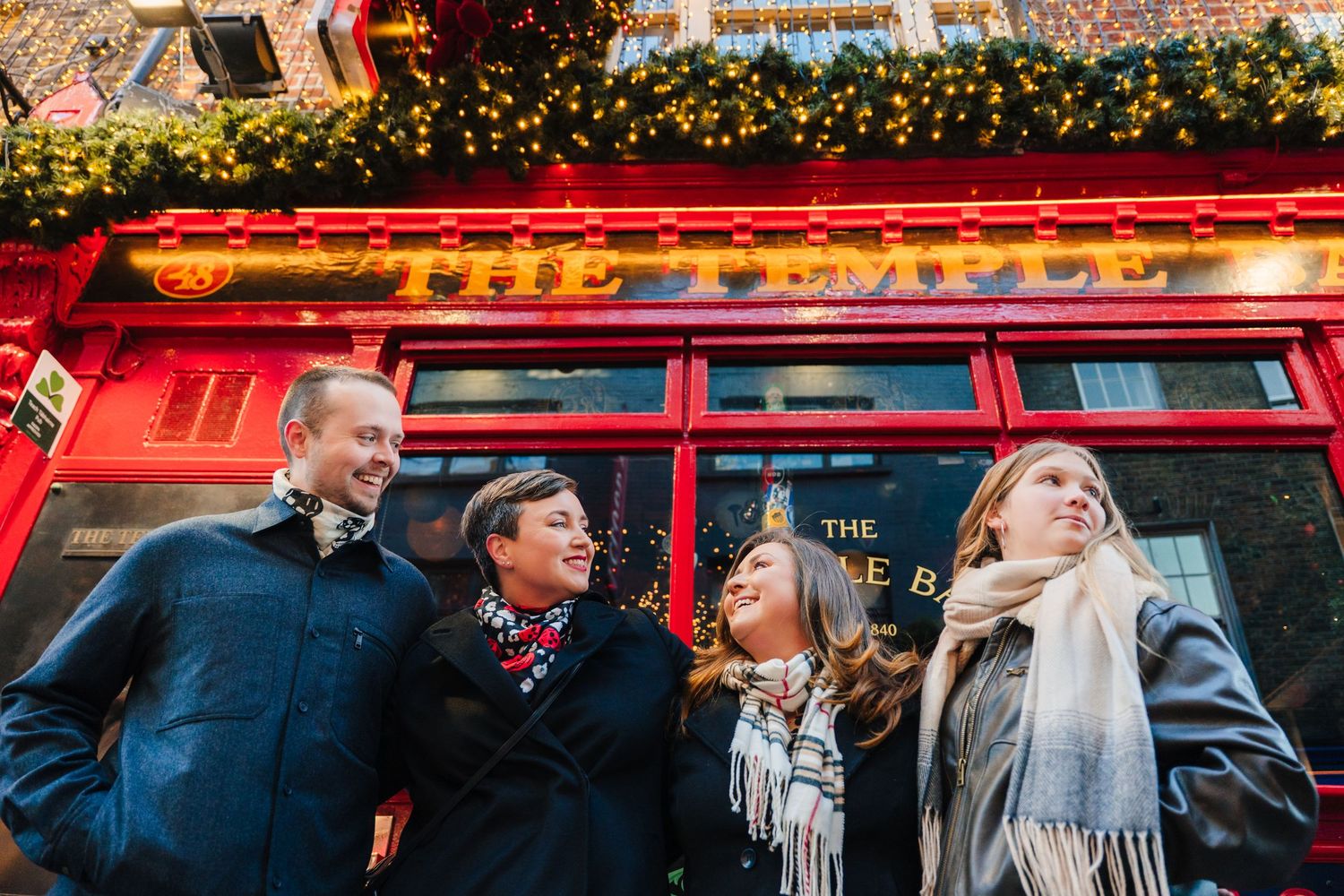 Festive string lights illuminate the exterior of a red-painted building with outdoor dining tables. Family walking through Dublin city centr