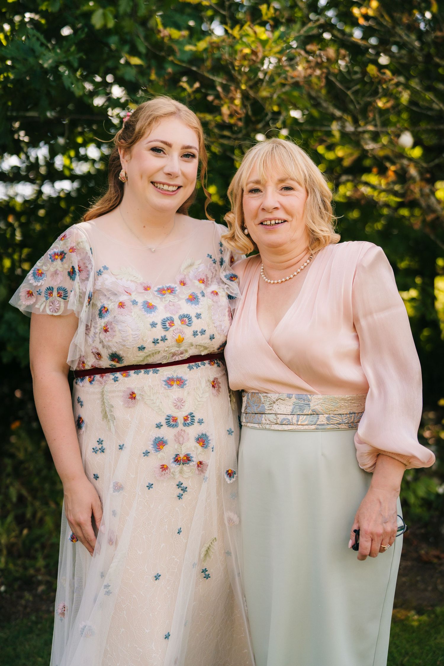 Two people wearing elegant dresses pose together outdoors, one in a floral pattern dress and one in a blush pink top with grey skirt. . Vill