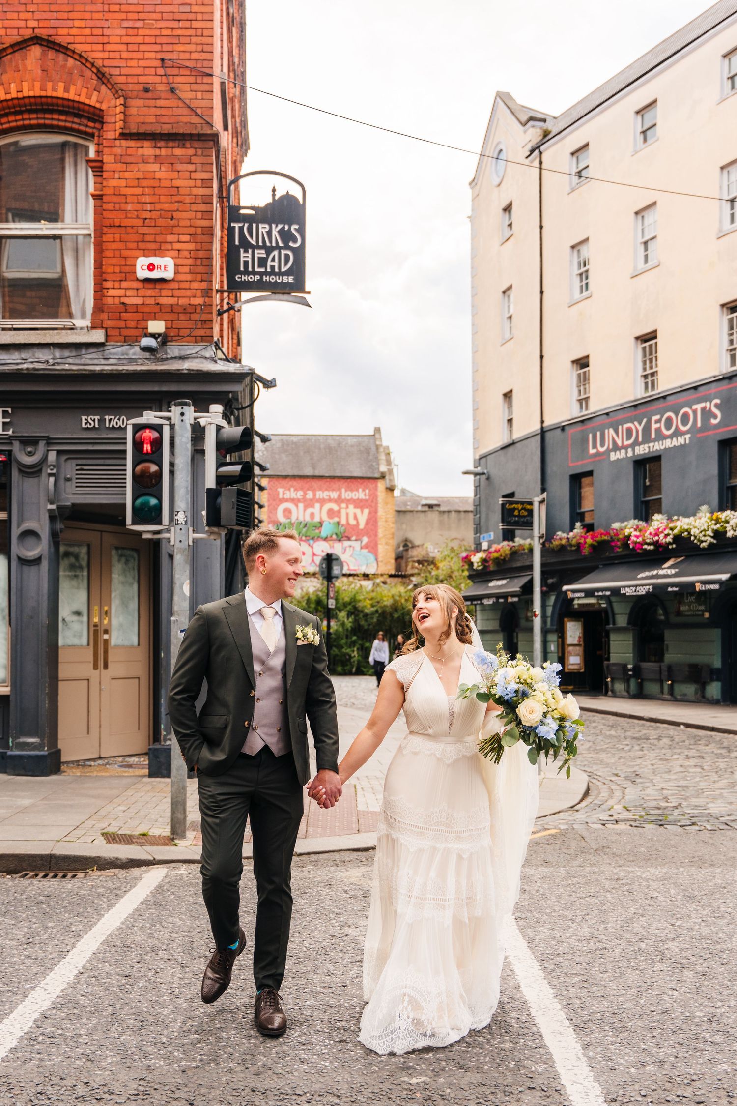 A couple in wedding attire walks down a cobblestone street past brick buildings and storefronts in an urban setting. PHOTOGRAPHER Dublin Ire