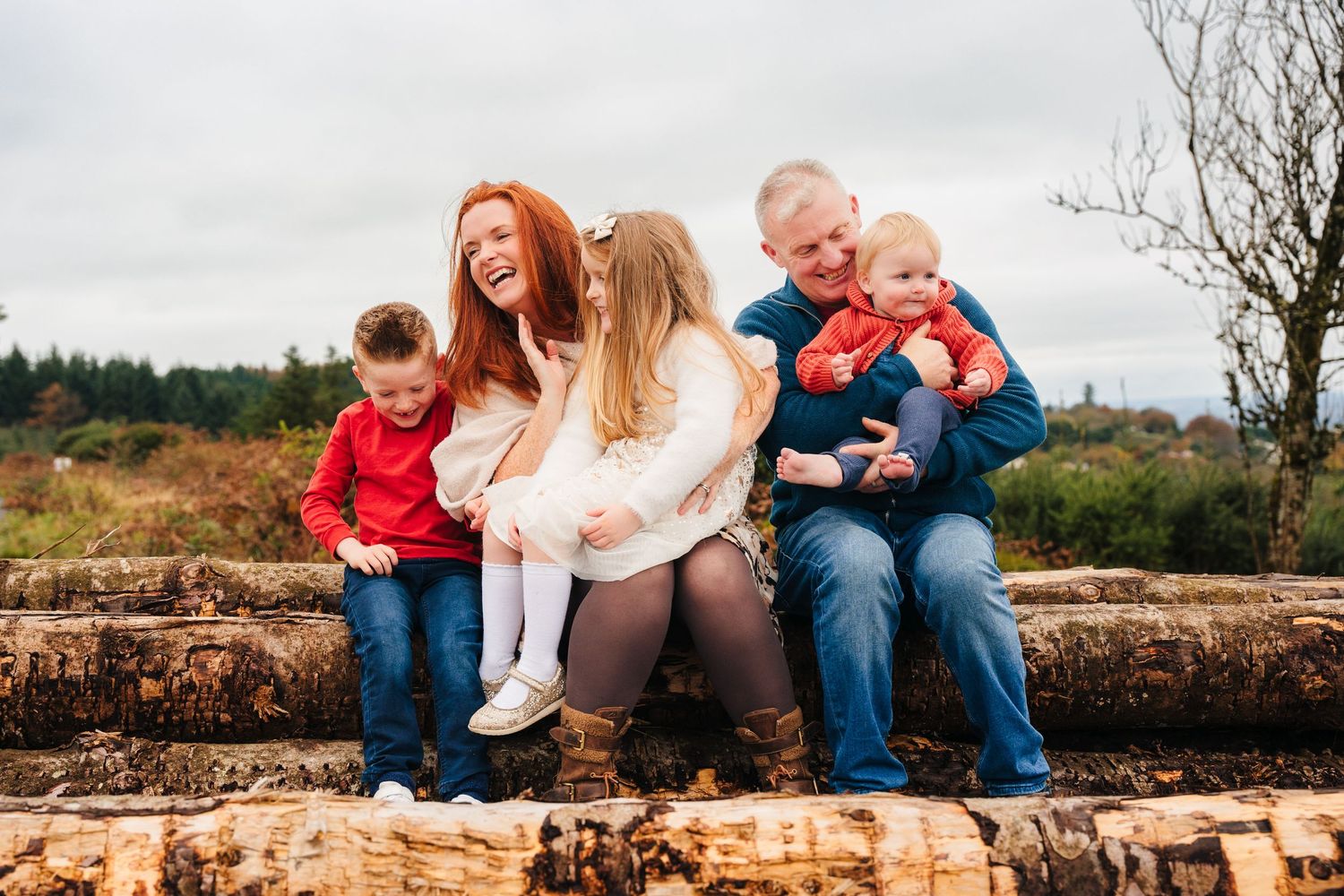 Family Photographer Dublin Ireland A family sits together on a rustic stone wall surrounded by autumn foliage in an outdoor setting.
