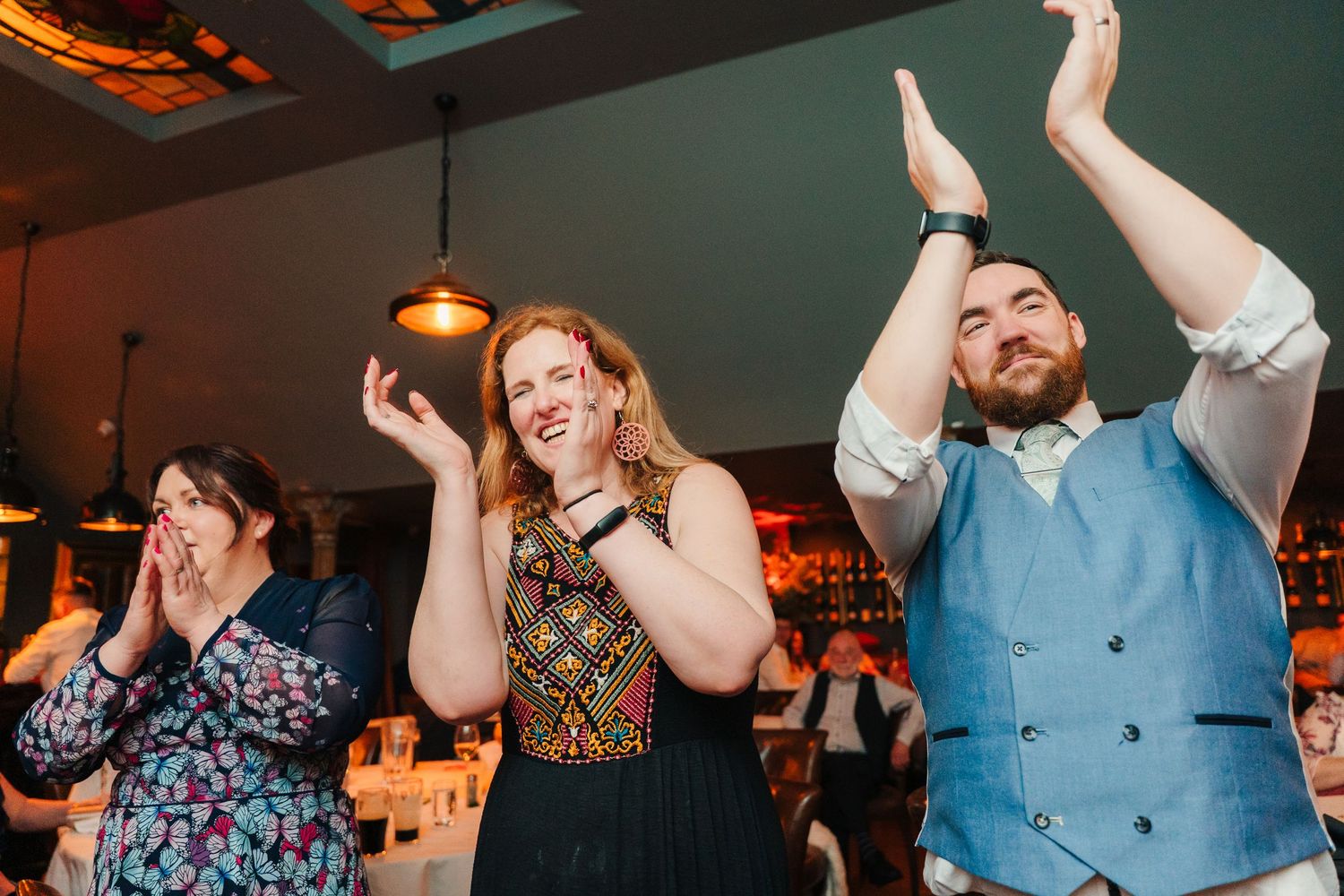 Wedding guests dancing and celebrating with enthusiastic arm gestures in a dimly lit venue. Marco Pierre Dublin Wedding Photographer