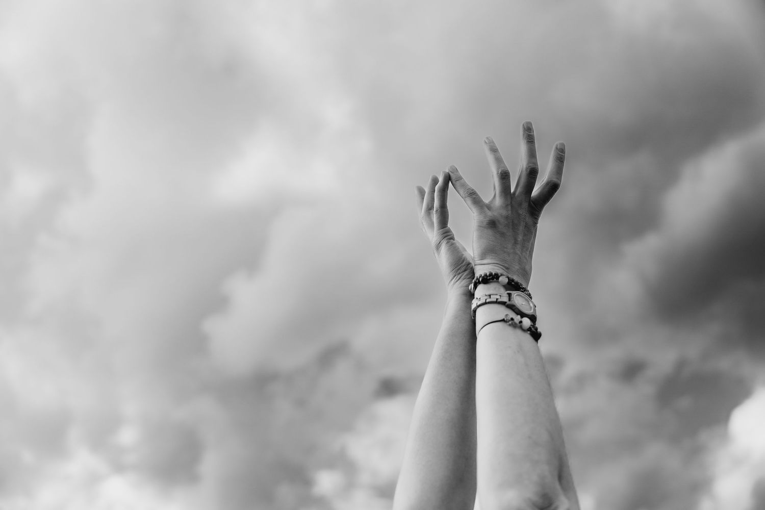 A raised hand reaching toward stormy clouds wearing multiple bracelets in dramatic black and white photography. Portrait PHOTOGRAPHER Dublin
