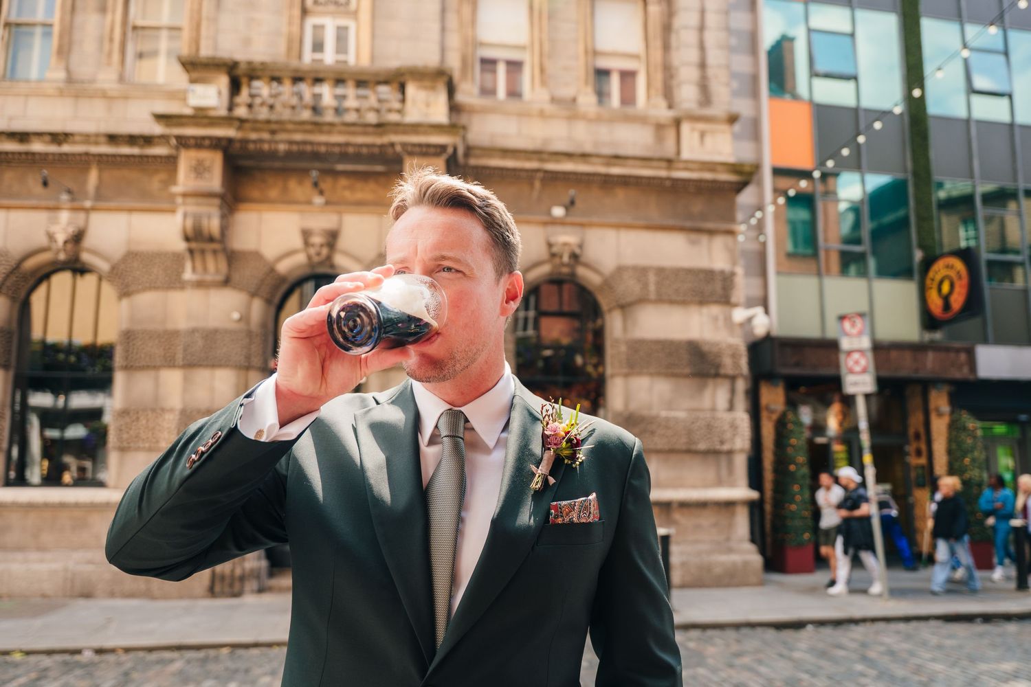 Well-dressed businessman in grey suit drinking coffee from disposable cup on city street with historic building facade. Dublin wedding photo