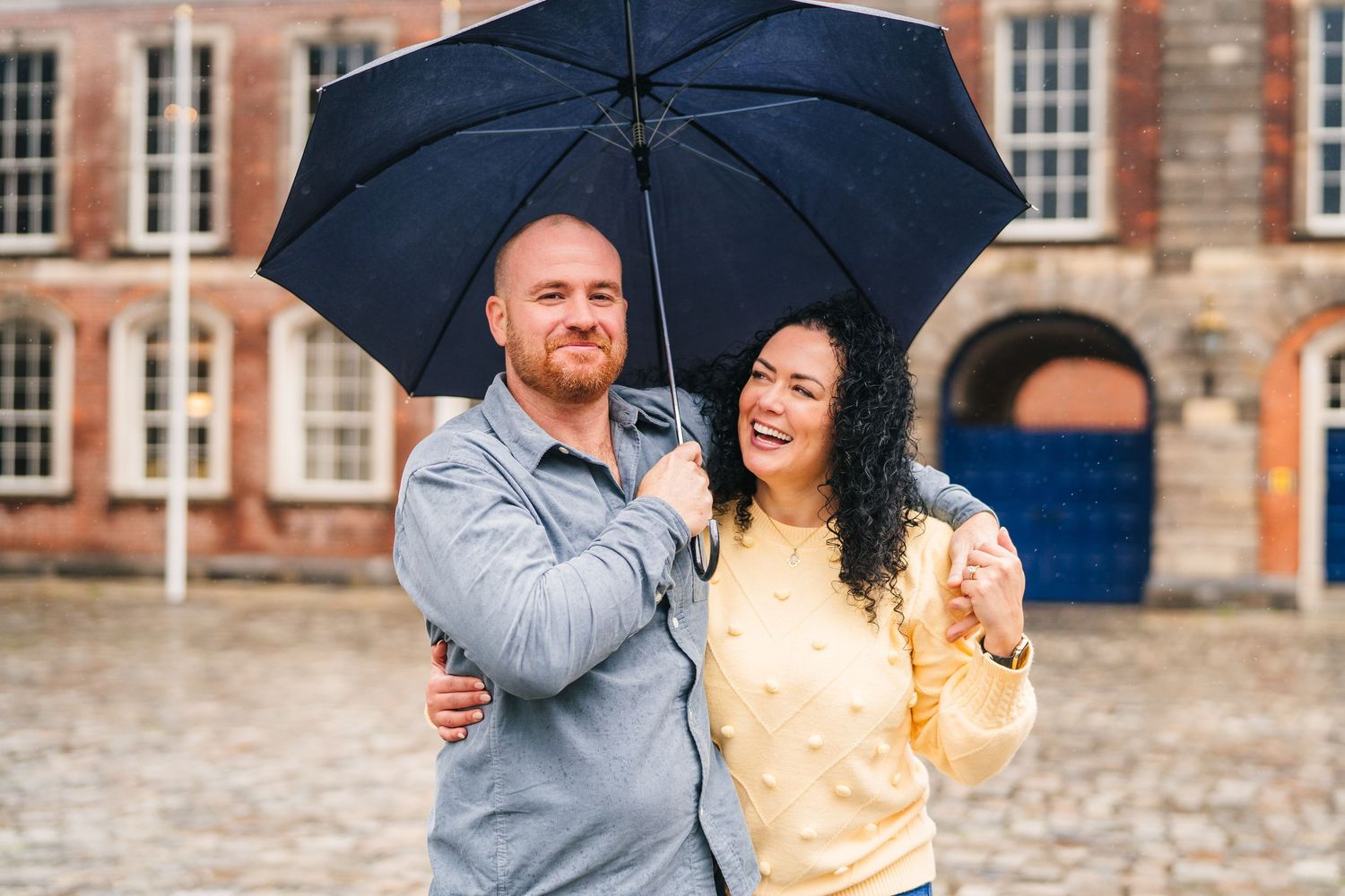 Two people share an umbrella while laughing together during a rainy day in front of historic brick building. Proposal Dublin Photographer