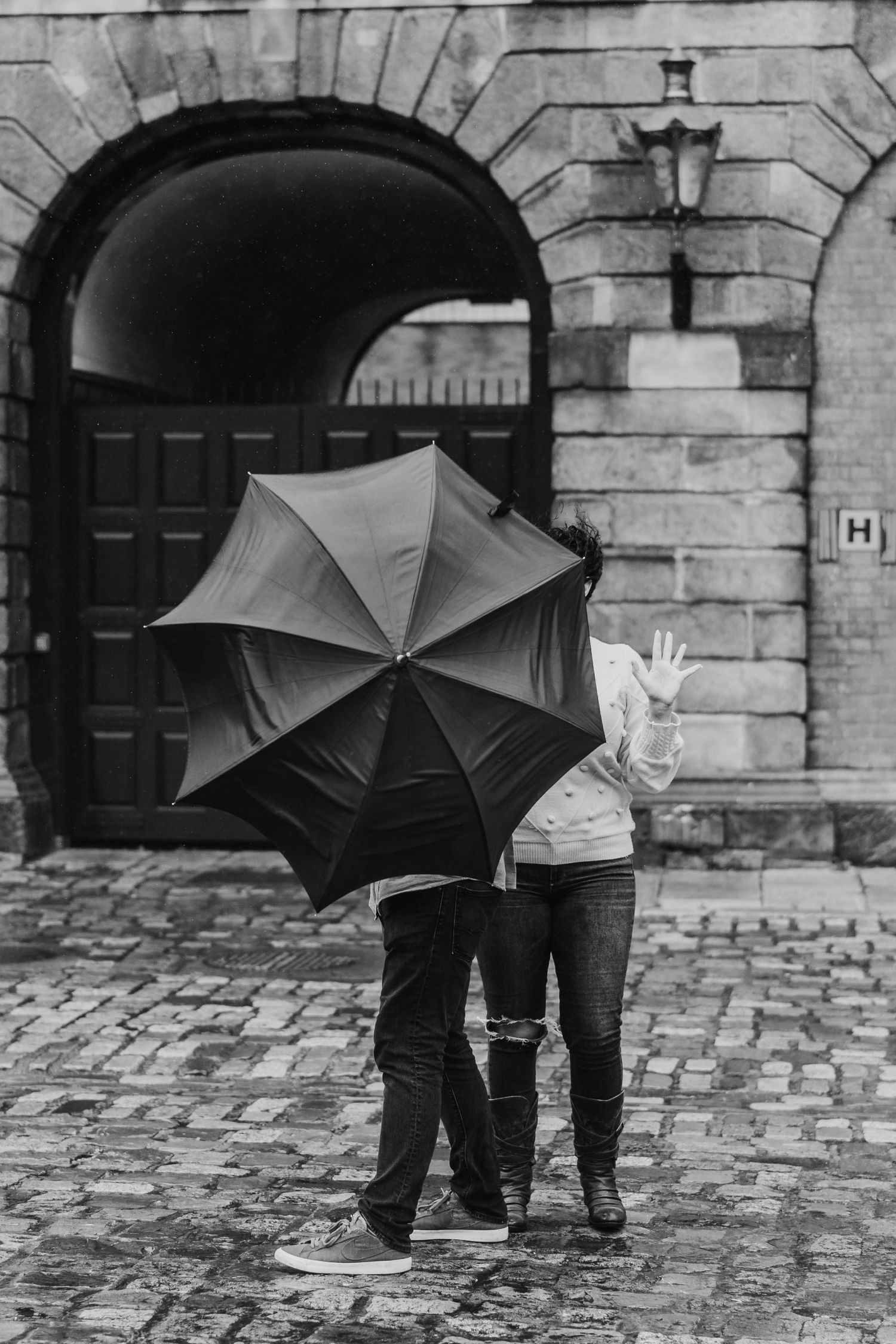 A black umbrella shields a couple standing on cobblestone street near an arched stone entrance in black and white. Proposal Dublin Photograp