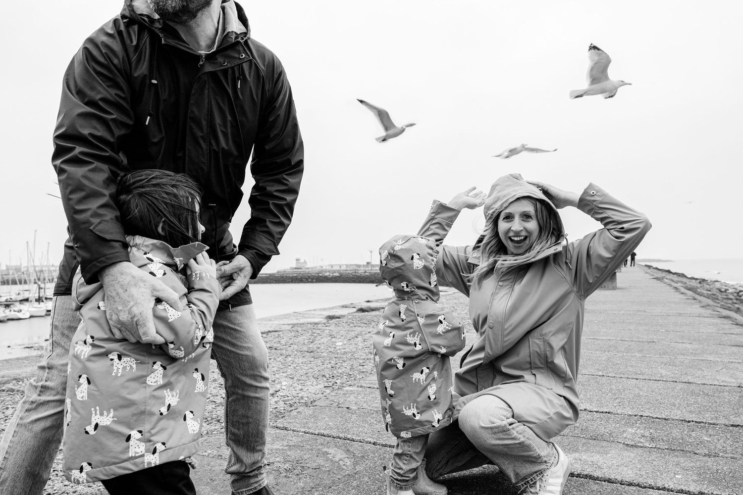 Winter-clad figures ducking and laughing as seagulls soar above them on a coastal path. PHOTOGRAPHER Dublin