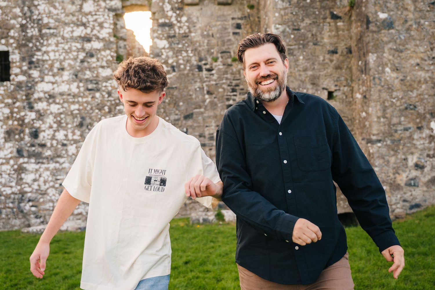 Two people laughing and walking together on grass against an old stone wall in casual attire. Photographer Ireland dublin