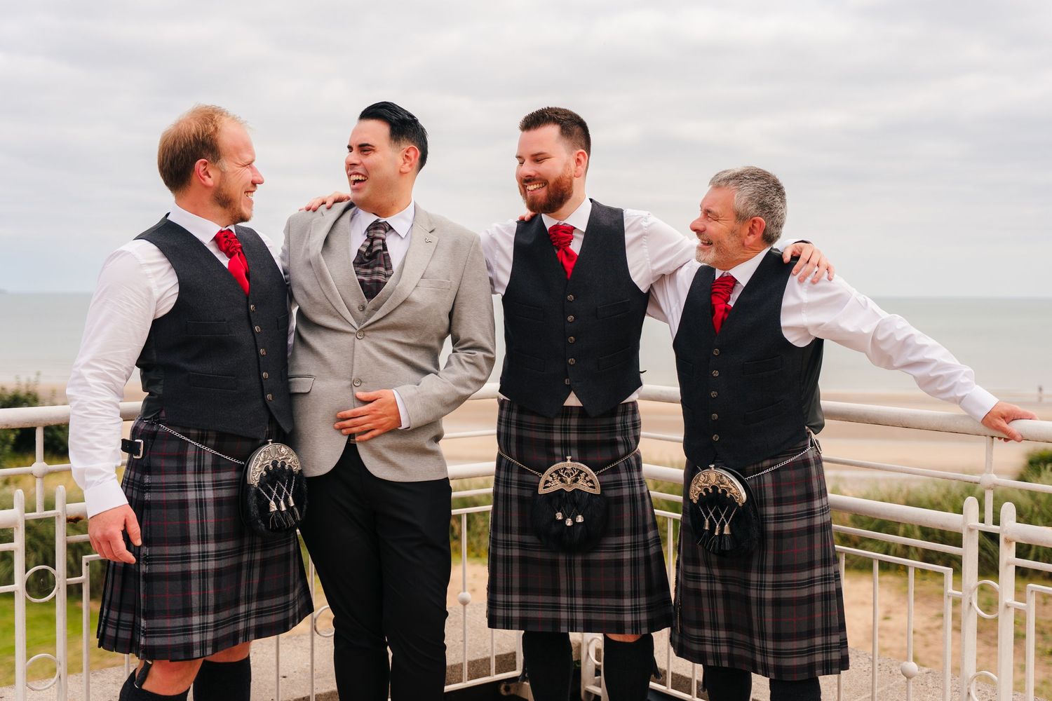 Groomsmen wearing traditional Scottish kilts share a candid laughing moment together at an outdoor wedding venue. Portmarnock Hotel Wedding