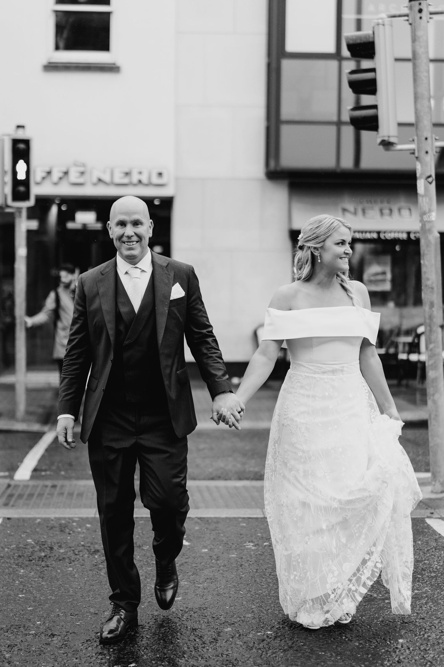 Black and white photo of a couple in wedding attire walking hand in hand down a city street. The Dean Dublin Wedding Photographer