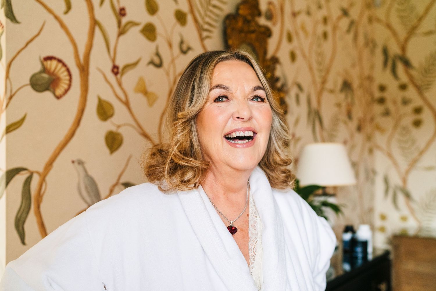 Person in white robe laughing joyfully against decorative floral wallpaper background. Virginia Park Lodge Wedding Photographer Dublin Meath