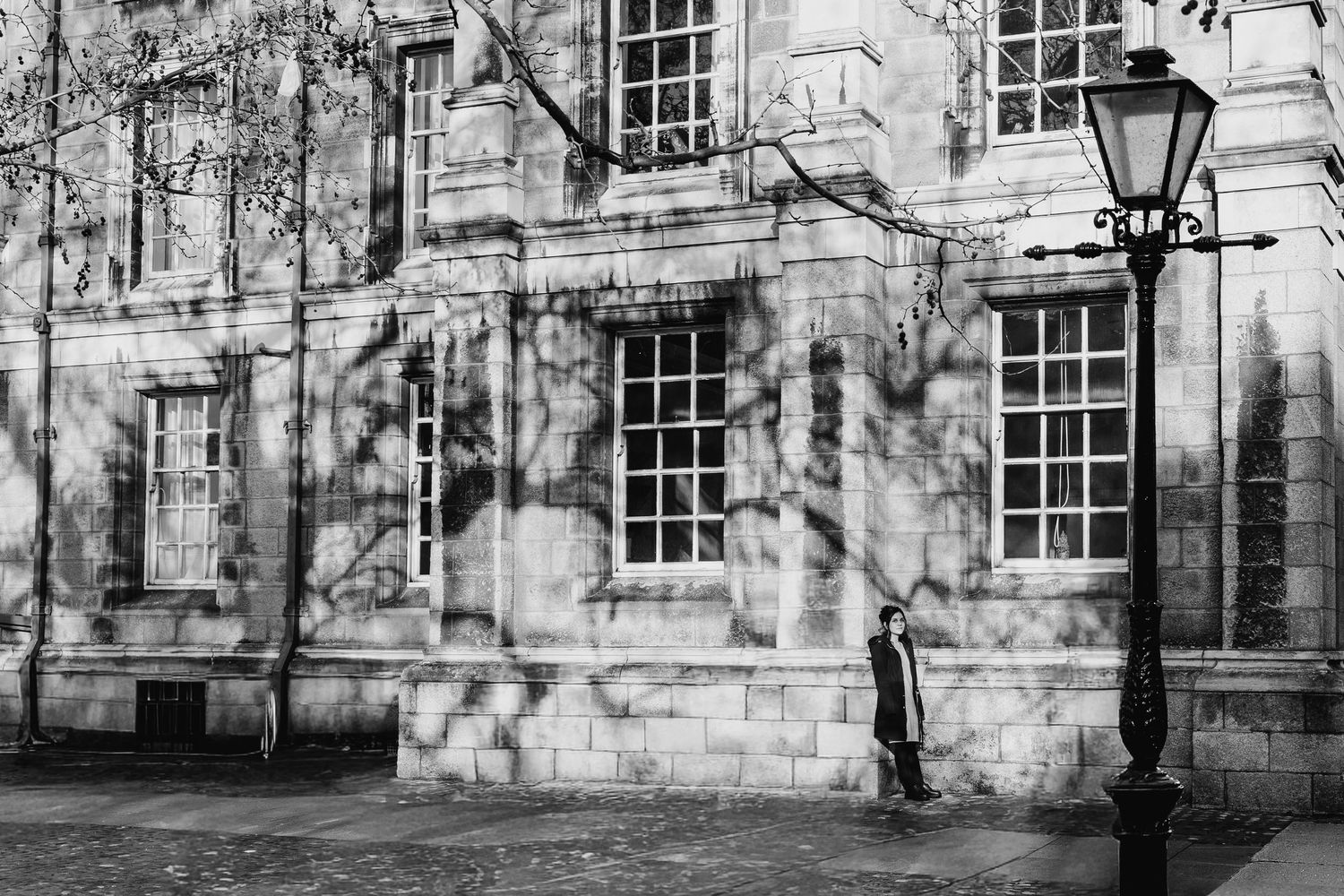 A person stands beside a historic stone building with tall windows and a vintage lamp post casting shadows in black and white. professional