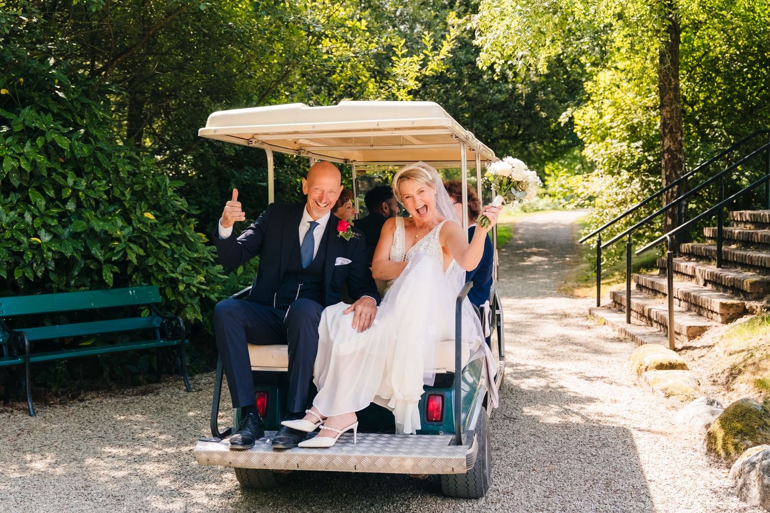 Wedding photographer Dublin Bride and groom in wedding attire giving thumbs up while riding a golf cart on a sunny outdoor path.