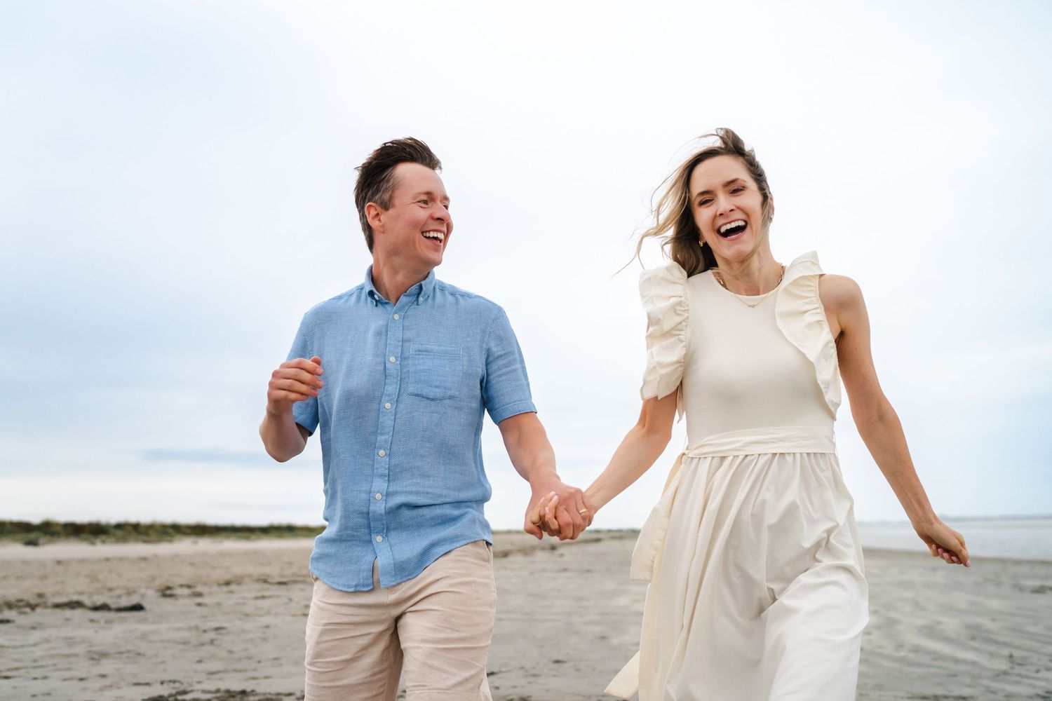 Couple laughing and running together along a scenic beach in casual summer outfits.  Family photographer Dublin Ireland