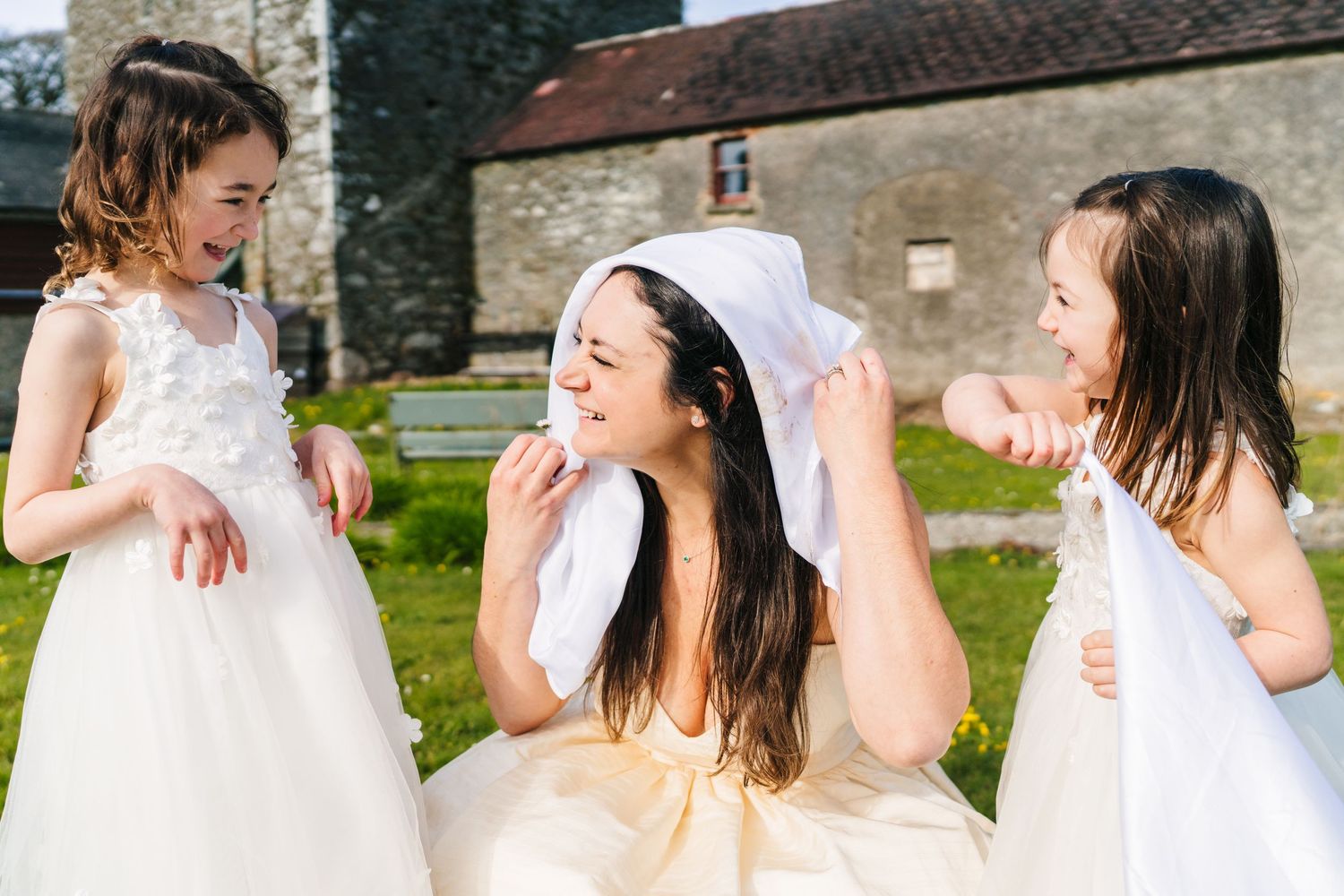 Three people in white dresses laugh and play together on a grassy lawn near stone buildings. Wedding PHOTOGRAPHER Dublin