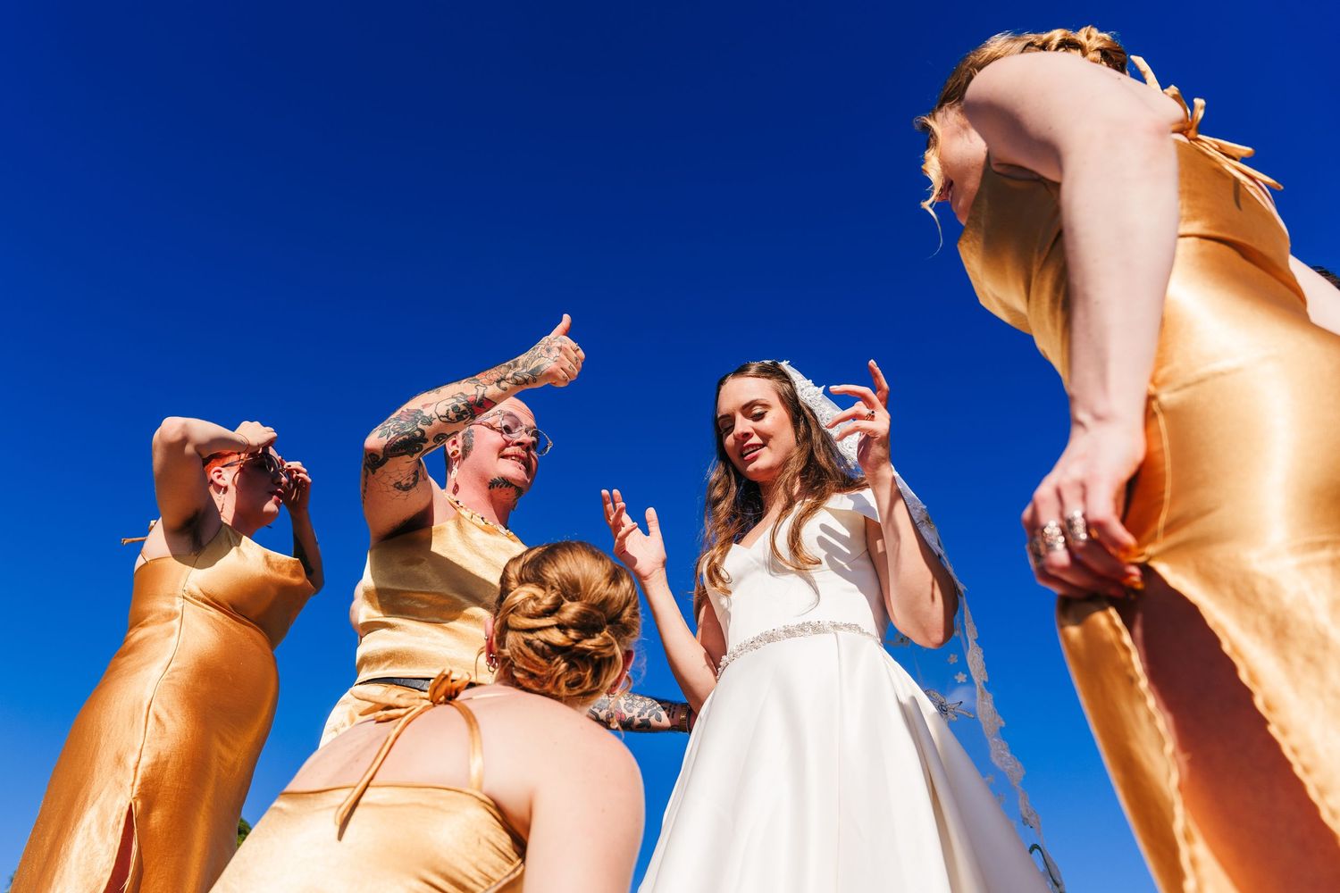 A wedding party in gold shimmering dresses stand together laughing against a bright blue sky. Wedding PHOTOGRAPHER Dublin