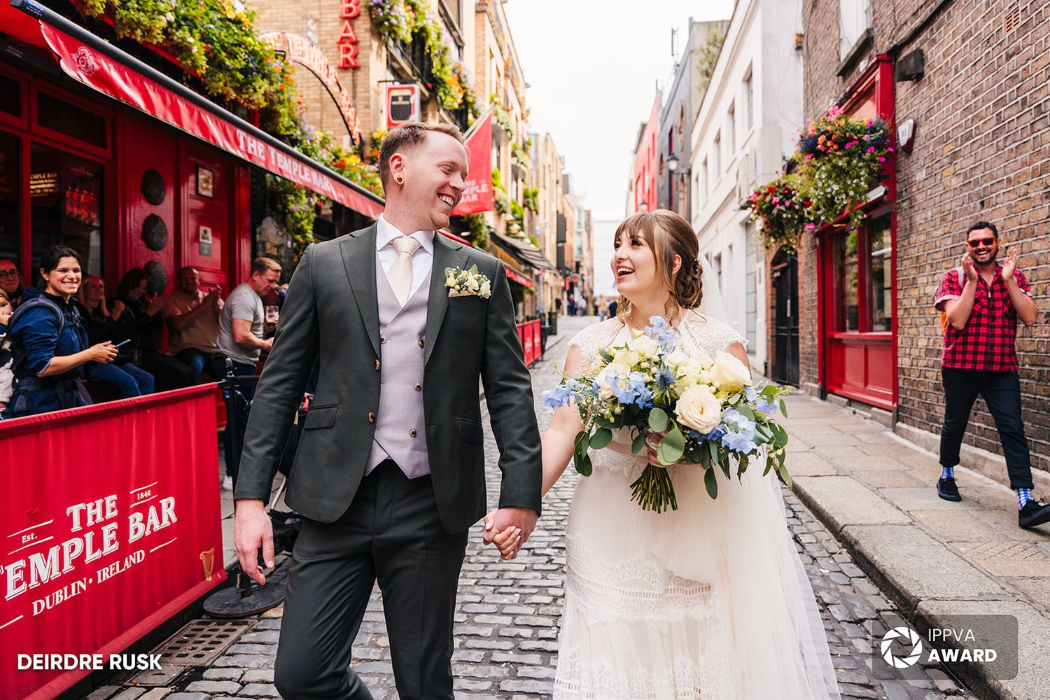 A wedding couple walks down a cobblestone street in Dublin with red storefronts and a photographer capturing the moment.