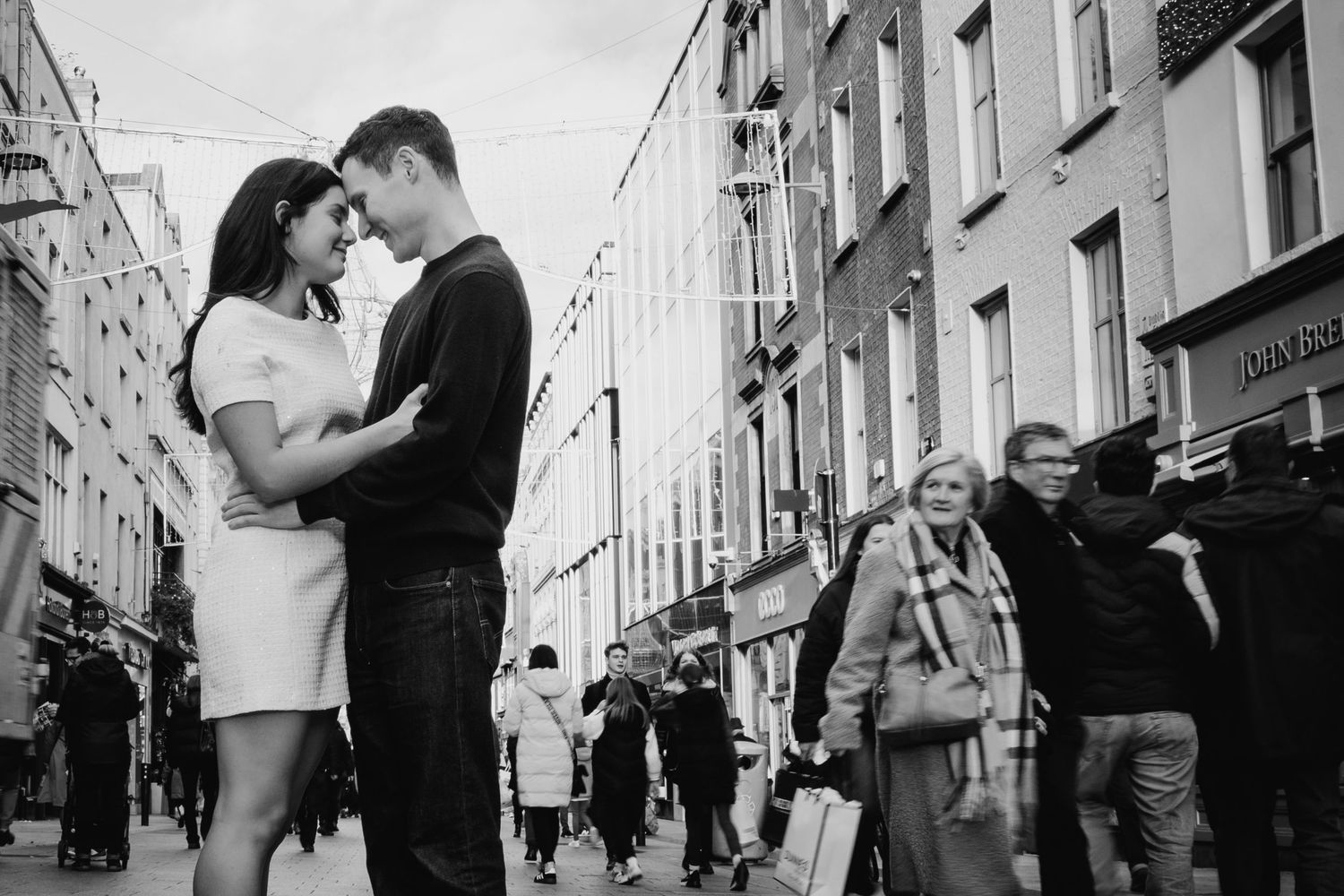 A romantic black and white street scene in an urban setting with pedestrians walking past buildings in the background. Surprise proposal dub