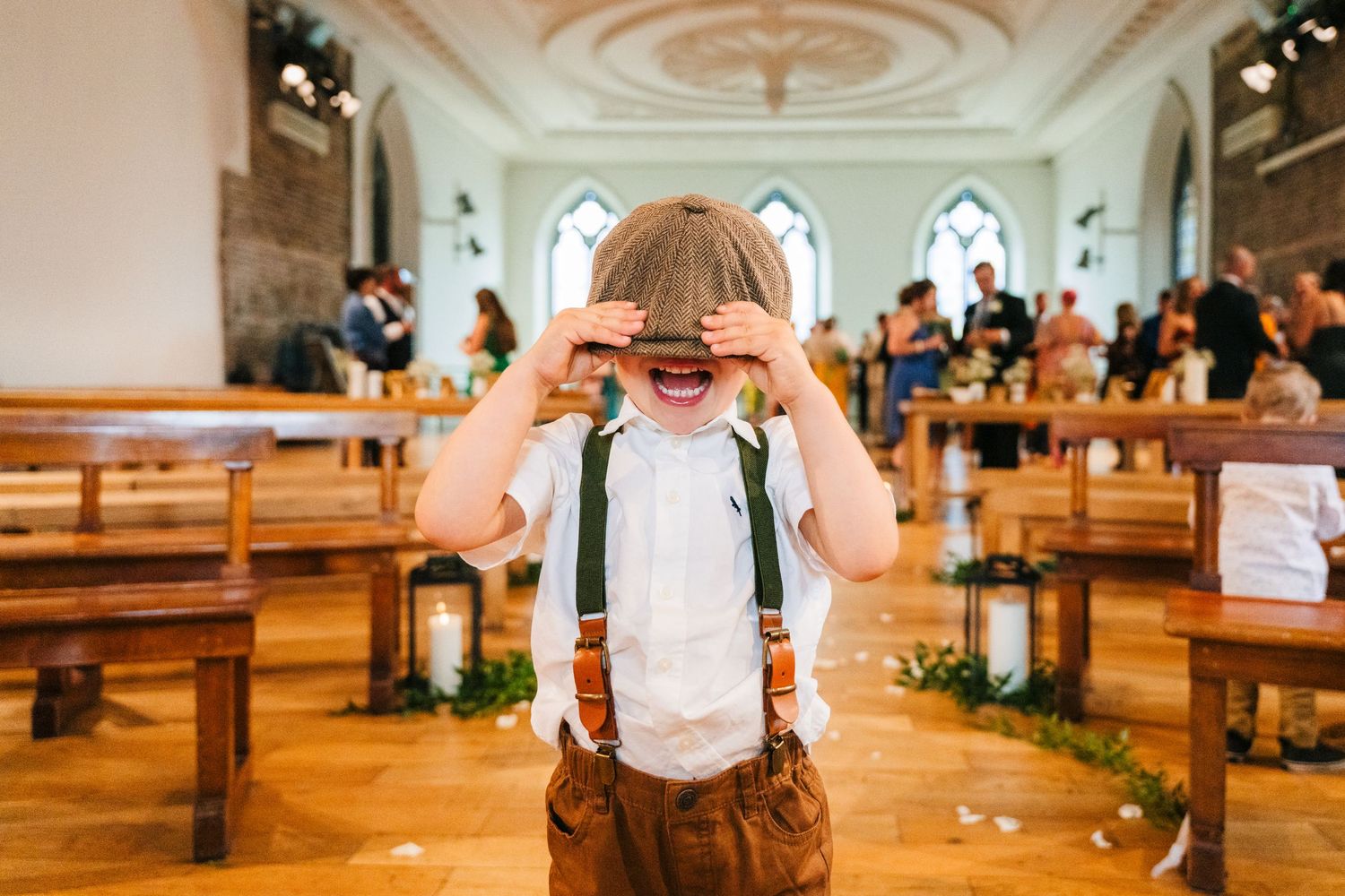 Child in suspenders and bow tie plays peek-a-boo in church during wedding celebration. PHOTOGRAPHER Dublin Ireland Wedding