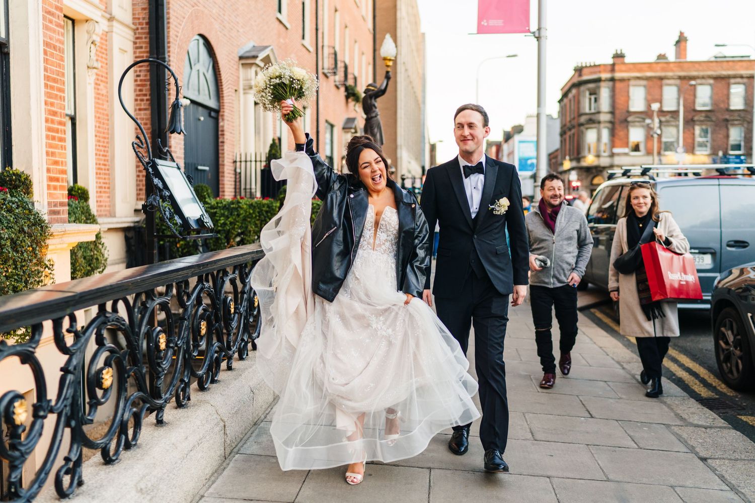 Newlyweds walk along a brick-lined street while guests follow behind. Wedding PHOTOGRAPHER Dublin
