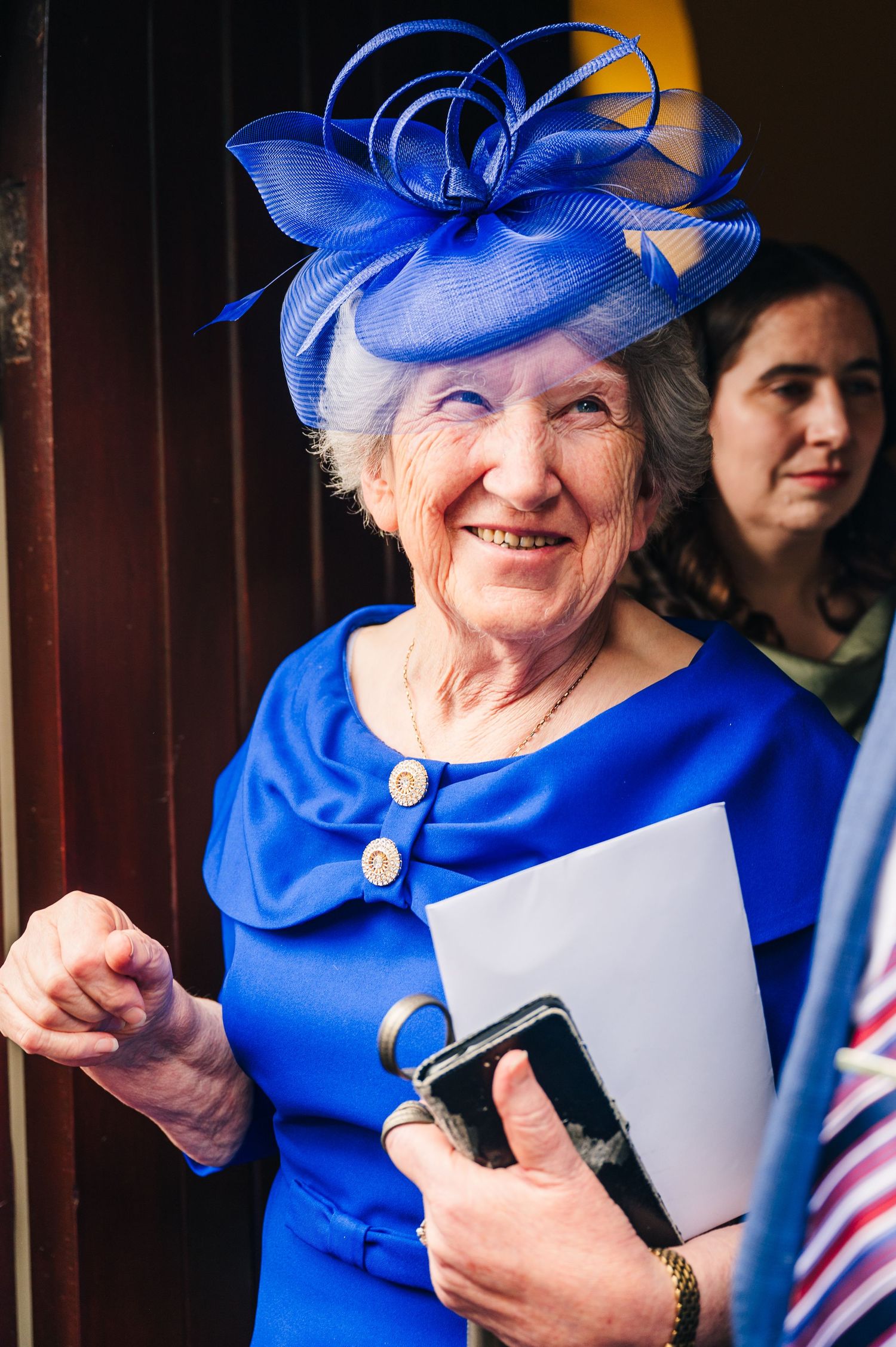 Millhouse slane wedding photographer dElegant woman in royal blue dress and matching fascinator hat smiles while holding papers and a phone.